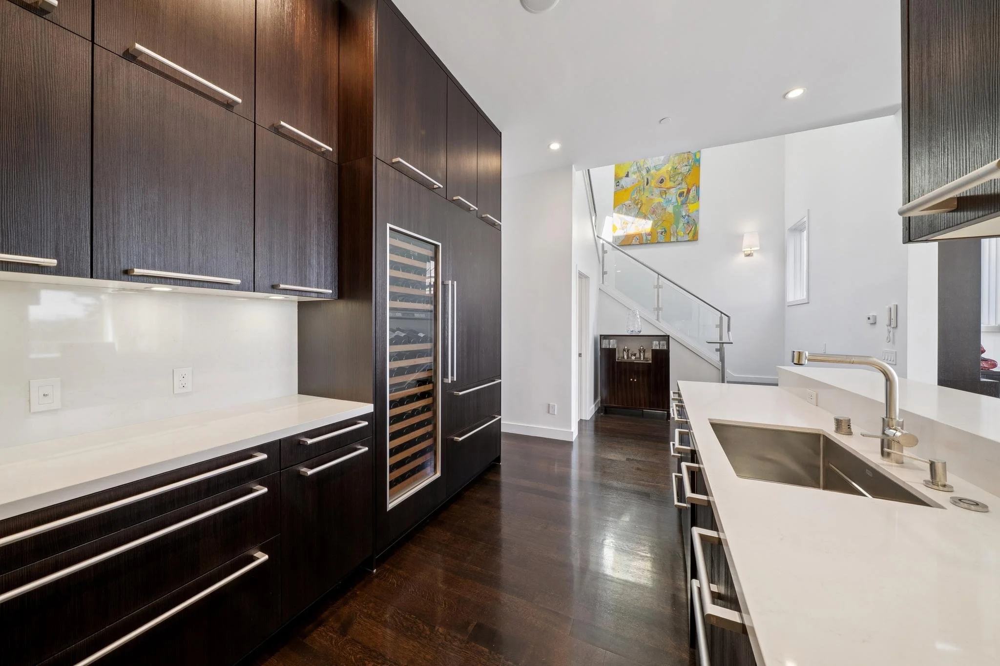 Modern kitchen with dark wood cabinets, white countertops, built-in wine cooler, stainless steel appliances, and hardwood floors. An open space with a staircase, abstract artwork, and large windows.