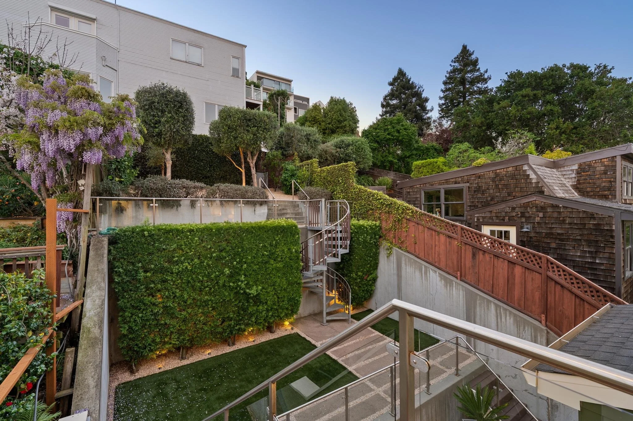 Multi-level backyard with a spiral staircase, green hedges, trees, and neighboring houses on a clear evening.