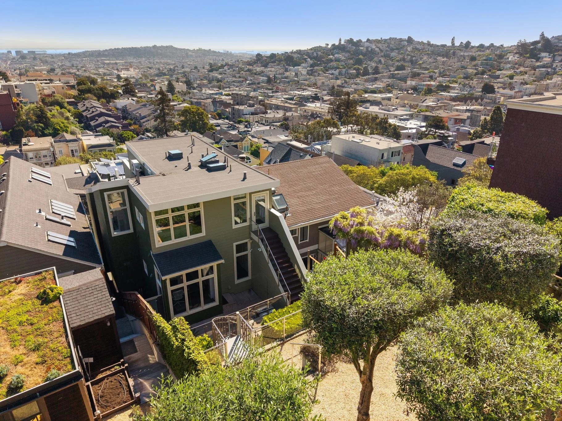 Aerial view of a city neighborhood with a modern green house, trees, and surrounding buildings under a clear sky.