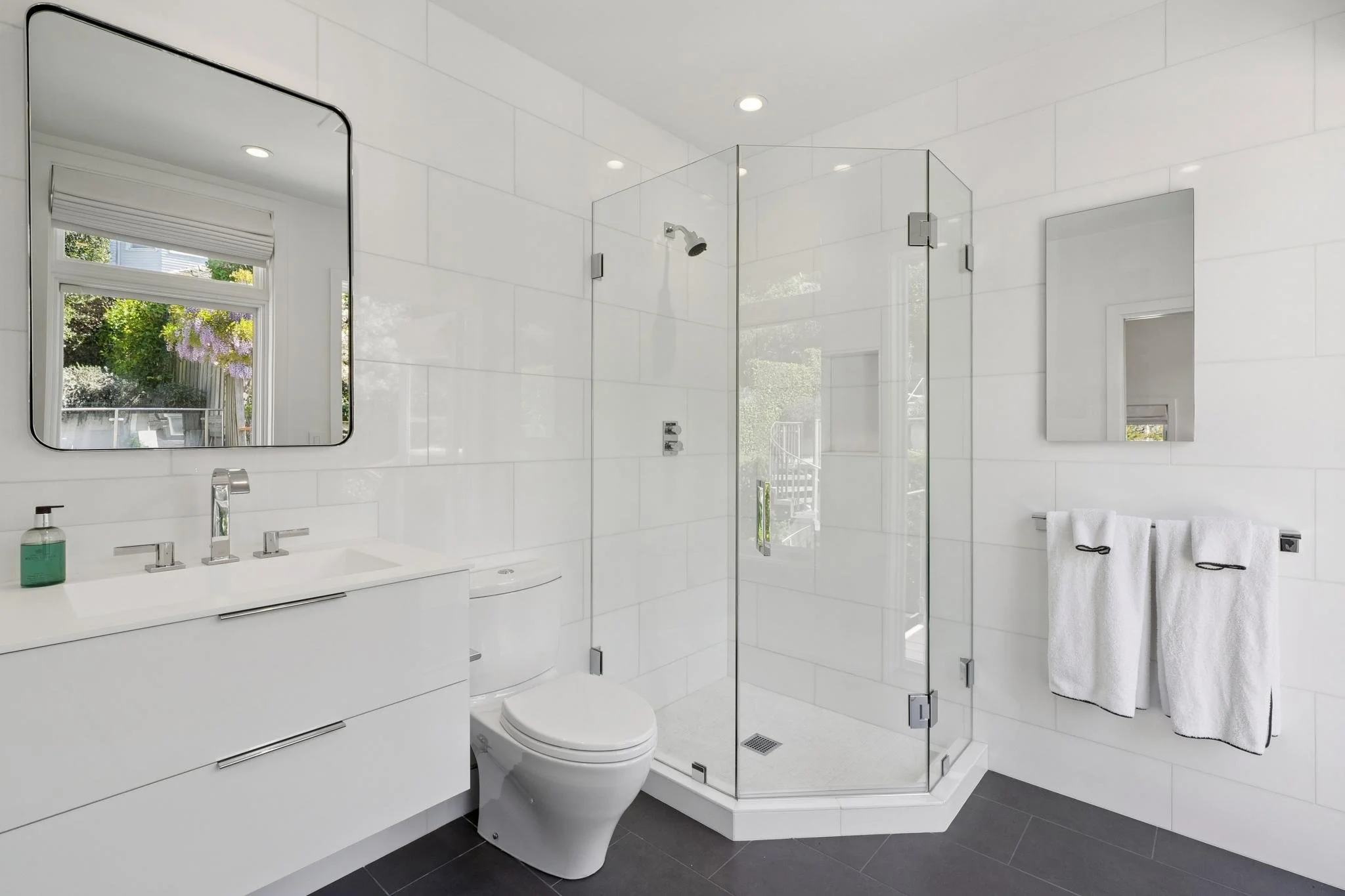 Modern white bathroom featuring a vanity with a mirror, a toilet, and a glass-enclosed shower with a window and towel rack.