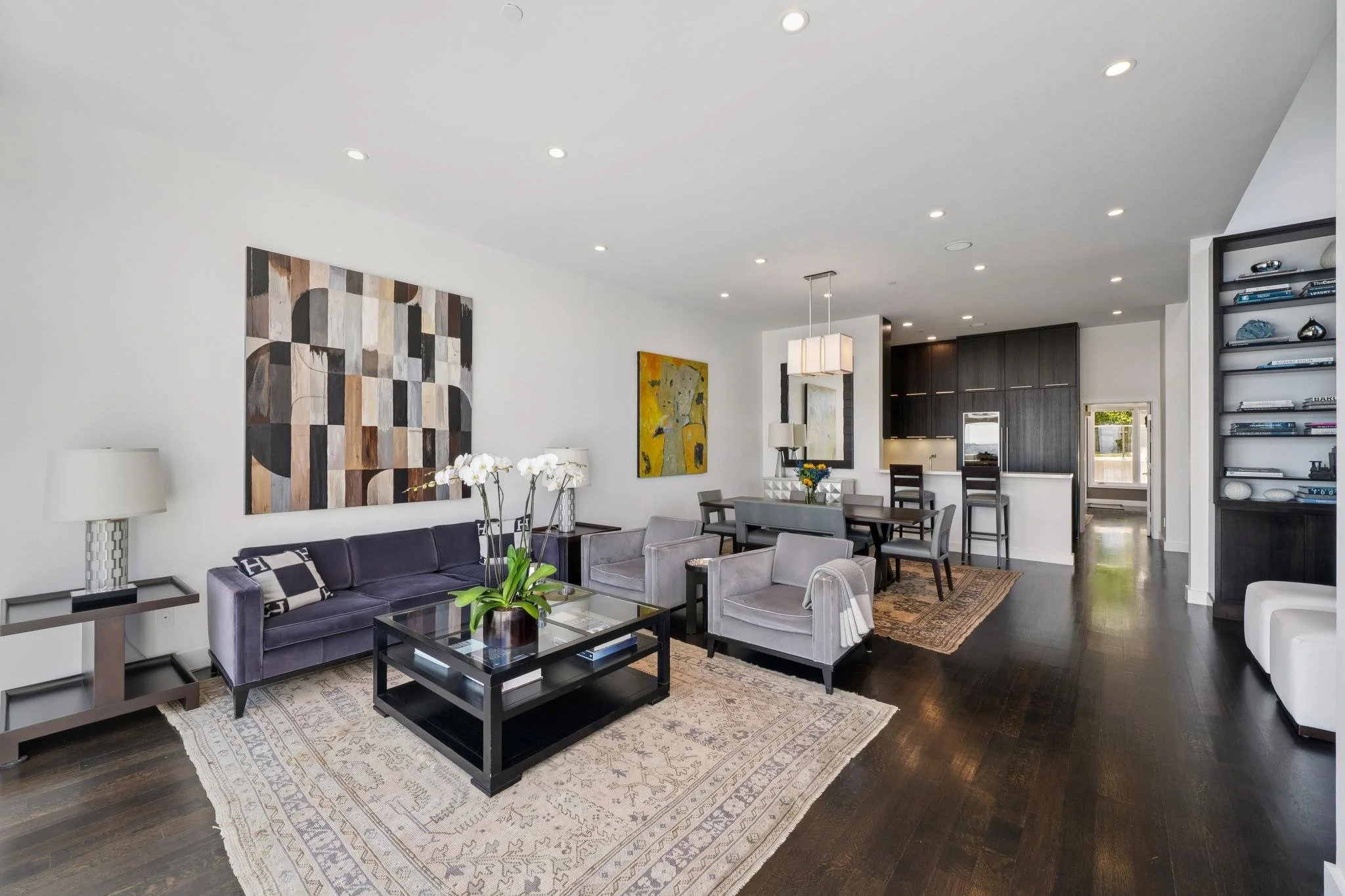 Living room with white walls, dark wood flooring, a purple velvet sofa, gray armchairs, a glass coffee table, and abstract artwork on the walls. Dining area with a table, chairs, and a chandelier in the background. Kitchen with dark cabinets and bar stools.