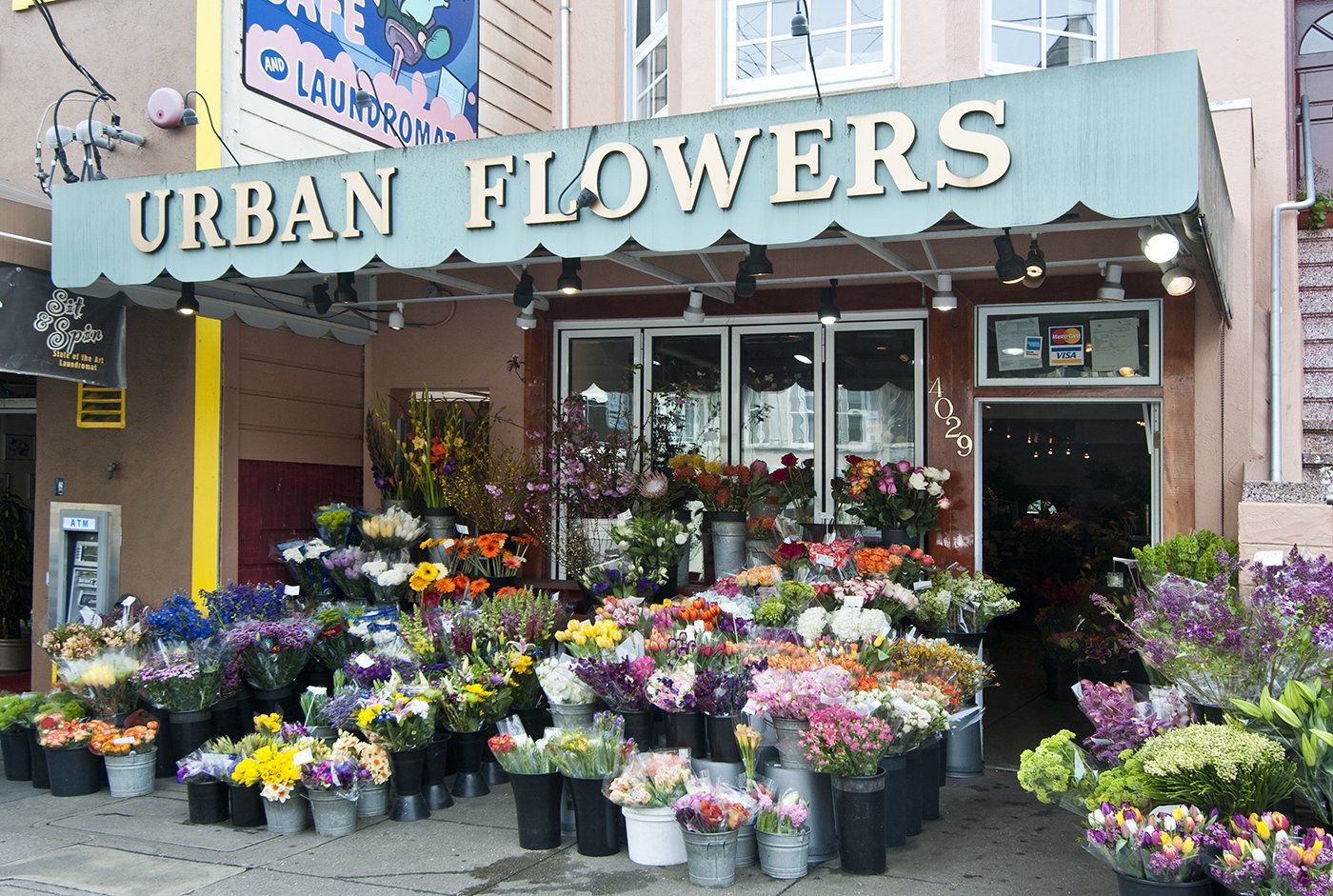 A flower shop named 'Urban Flowers' with an outdoor display of colorful flowers in buckets and vases, located on a city sidewalk in front of a storefront.
