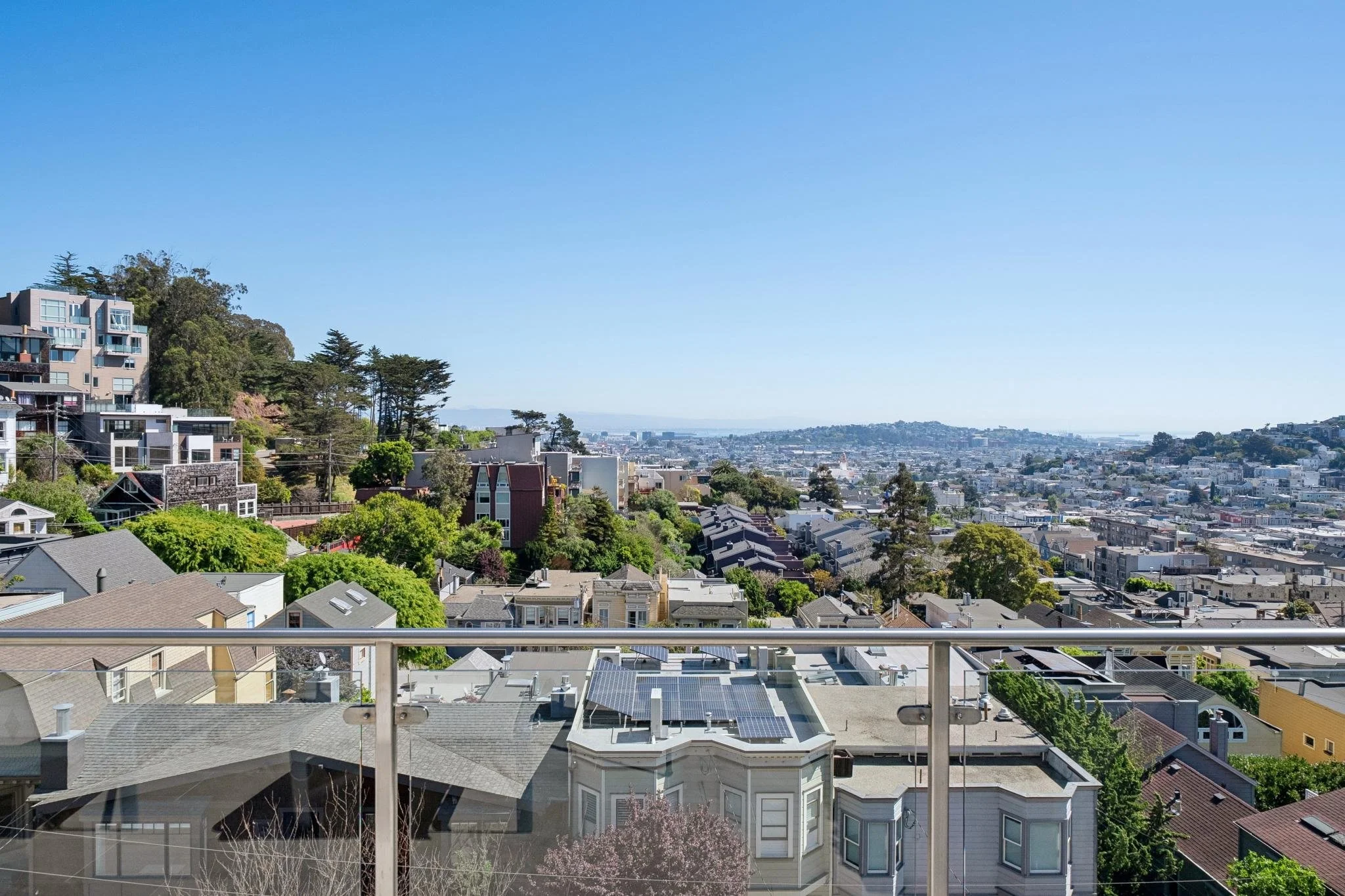 View from a balcony overlooking a city with residential houses, trees, and hills in the distance under a clear blue sky.