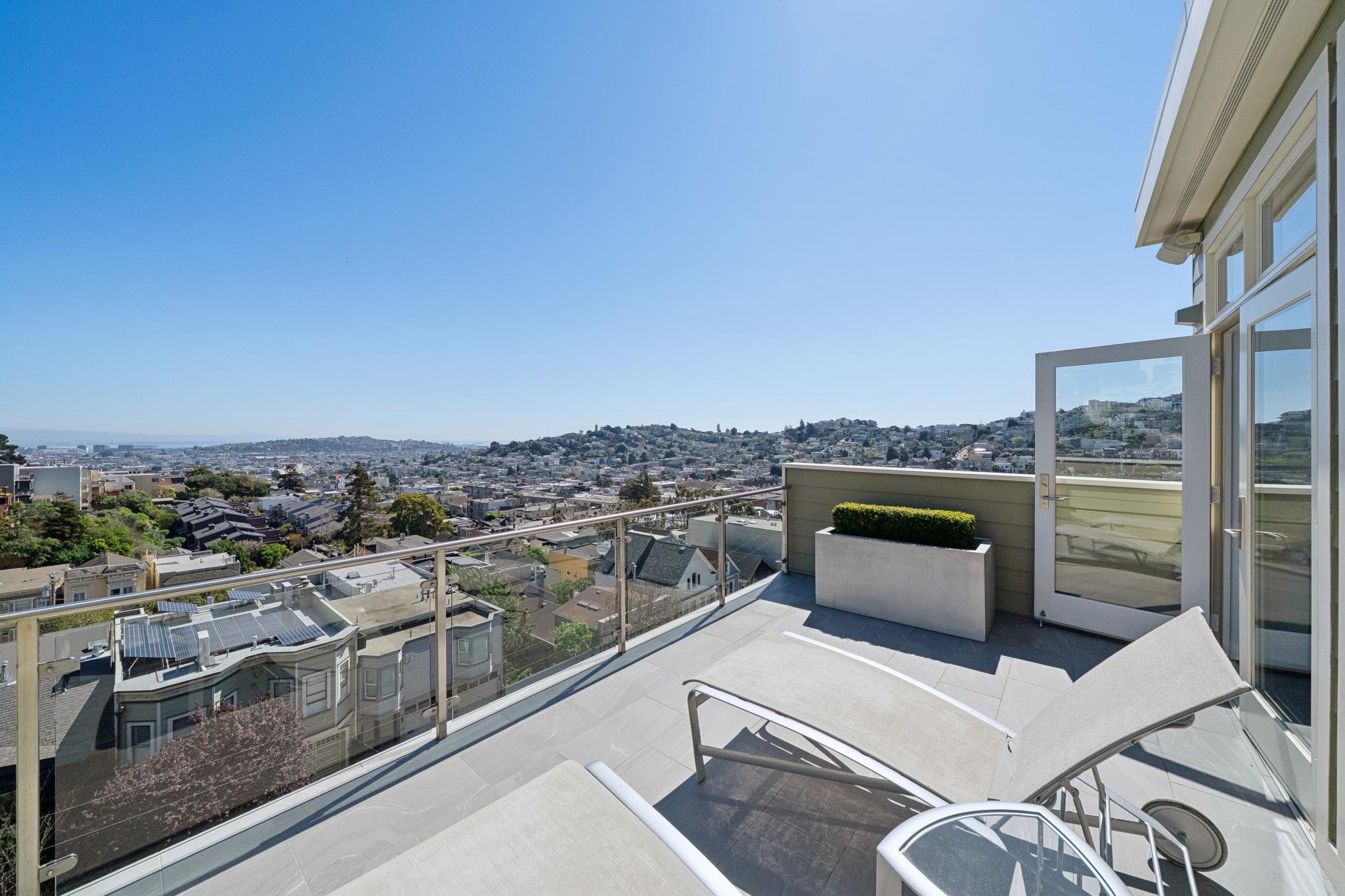 View from a balcony with glass railing overlooking a city and hills, sunny sky, lounge chairs, potted plant, open door.