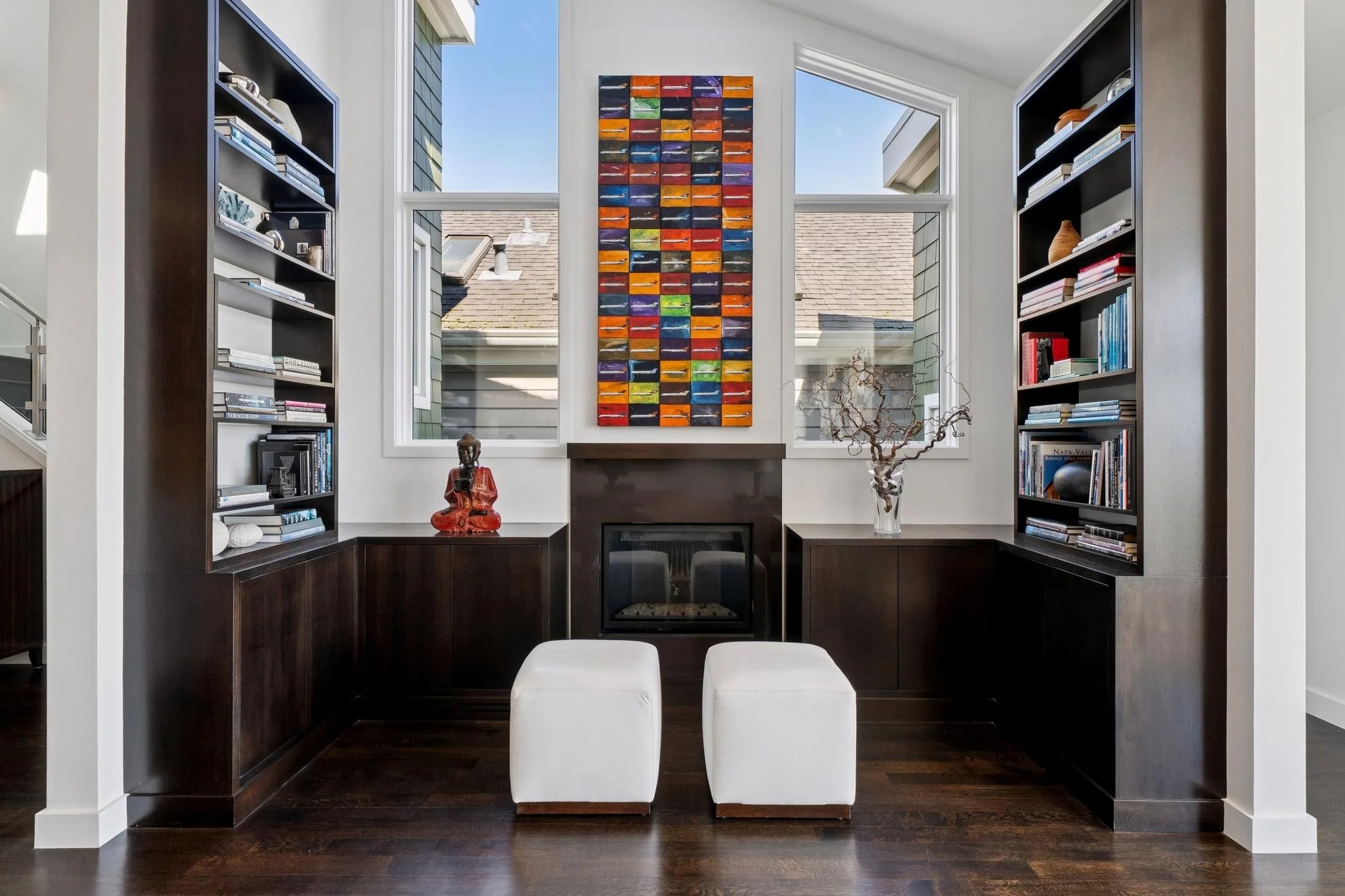 Living room with built-in dark wood bookshelves, a modern fireplace, artwork with colorful horizontal stripes, and two white ottomans. Large windows let in natural light.