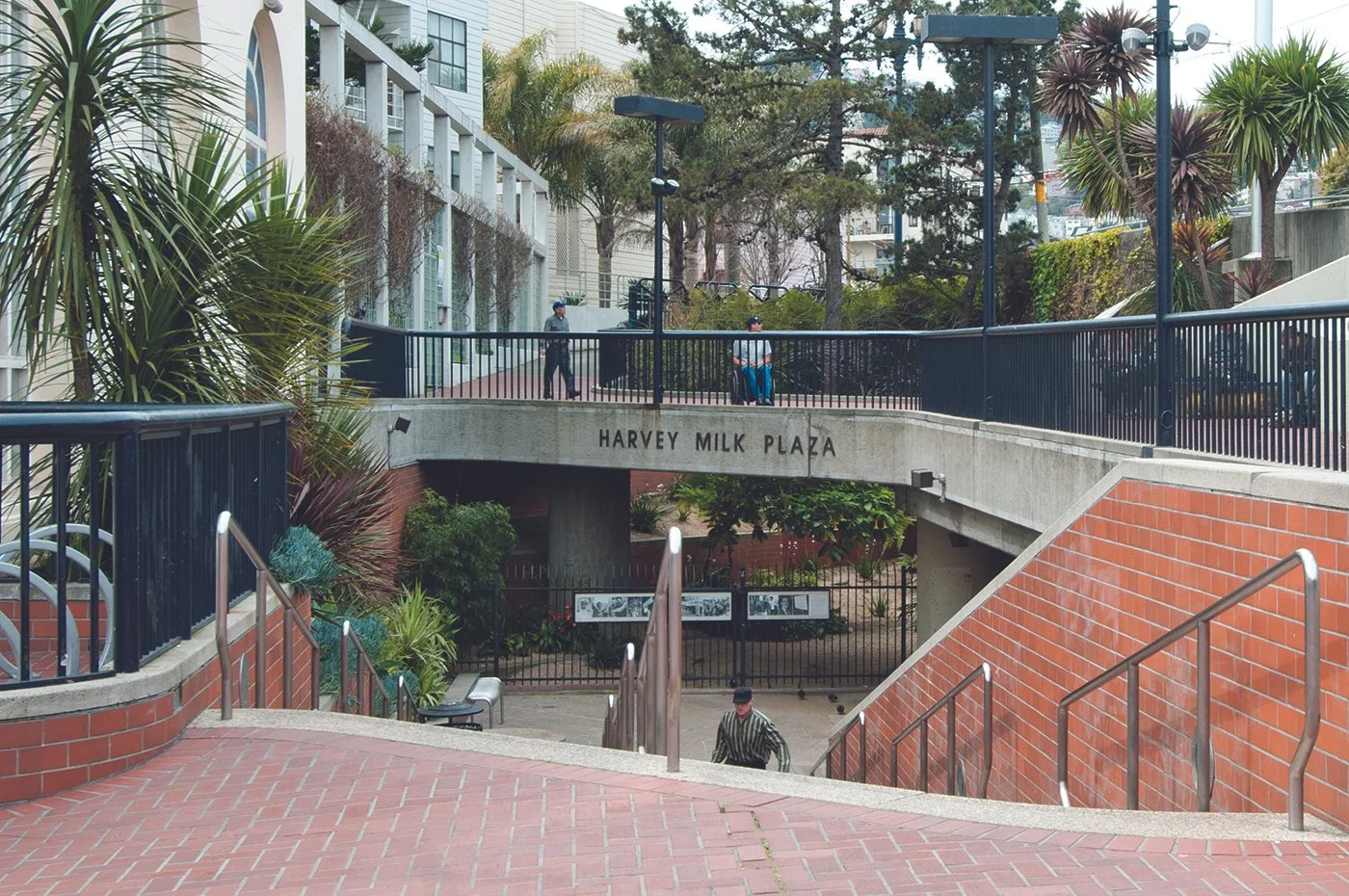 Public plaza with stairs, railings, and a sign that reads 'Harvey Milk Plaza.' People are walking and sitting, surrounded by trees and buildings.