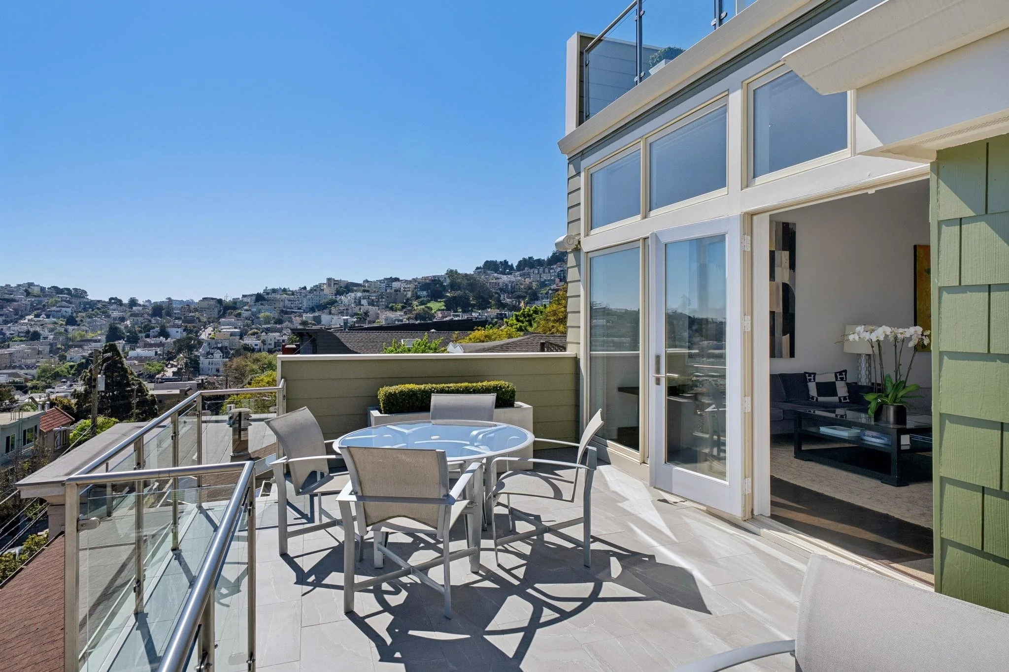Modern outdoor rooftop patio with a round table and chairs, overlooking a cityscape with numerous houses and hills in the distance.