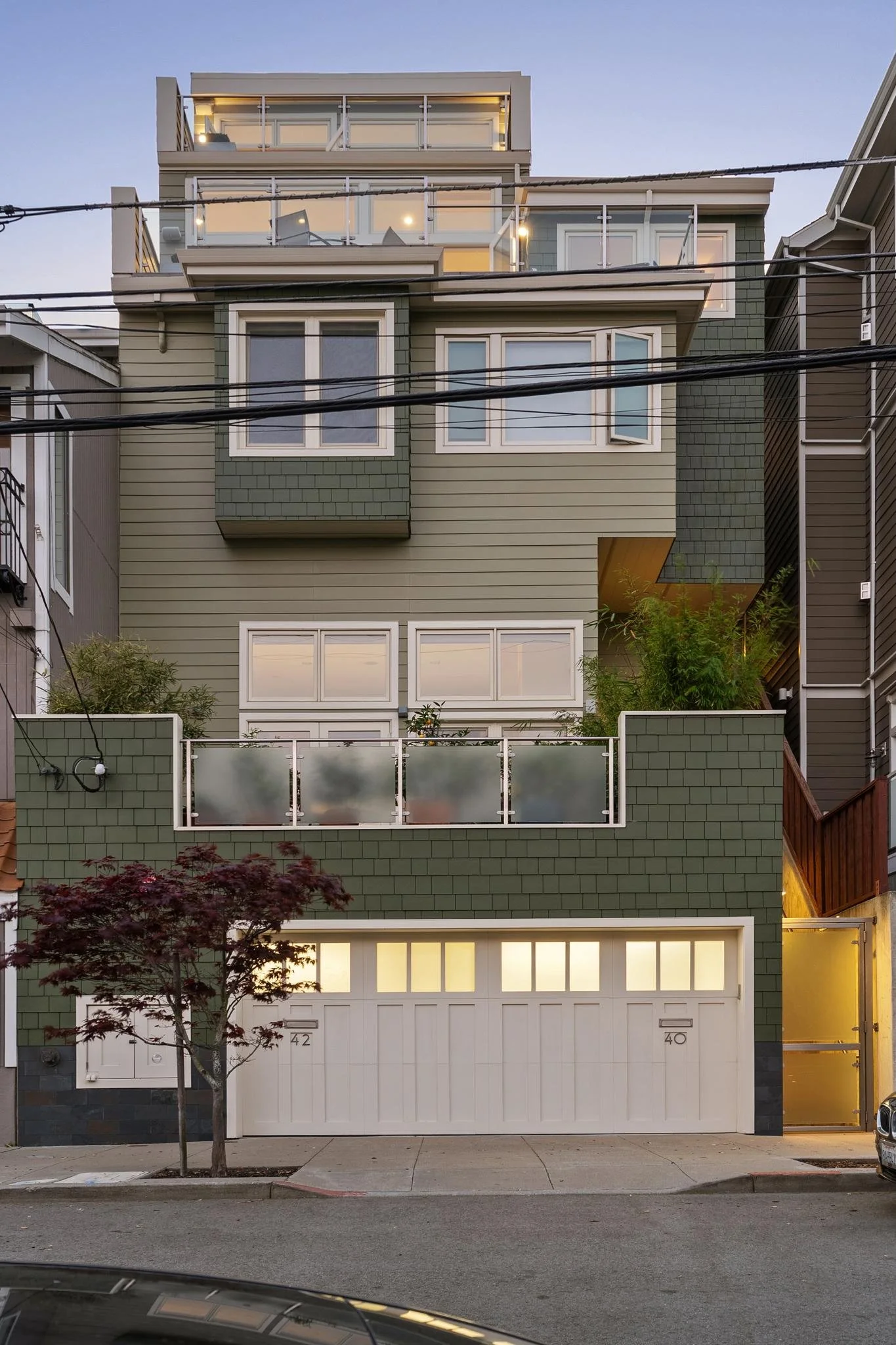 Multi-story modern house with a green exterior, white garage door, and large windows, located on a city street at dusk.