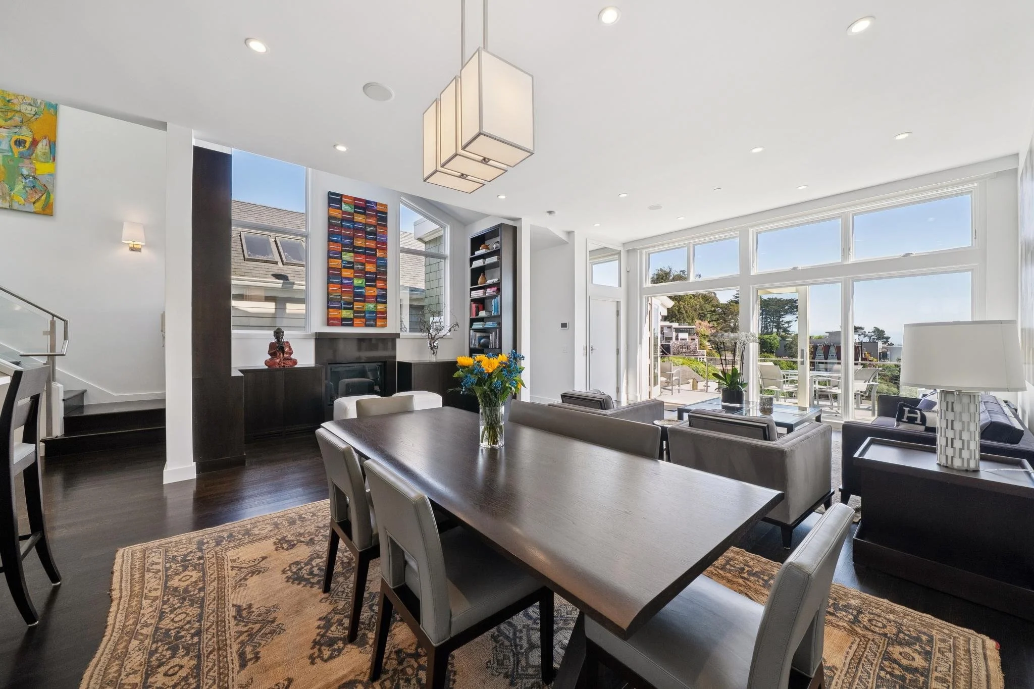 Modern living room with large windows, dark hardwood floors, a dining table with 8 chairs, a sitting area with sofas, coffee table, and outdoor patio visible through the windows.