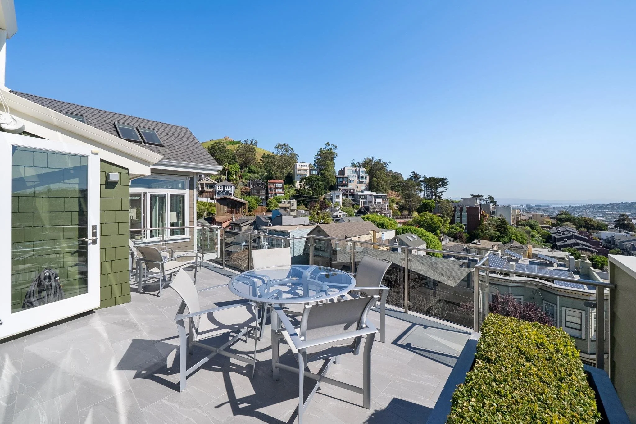 Rooftop terrace with outdoor table and chairs overlooking a hillside neighborhood with houses and greenery under a clear blue sky.