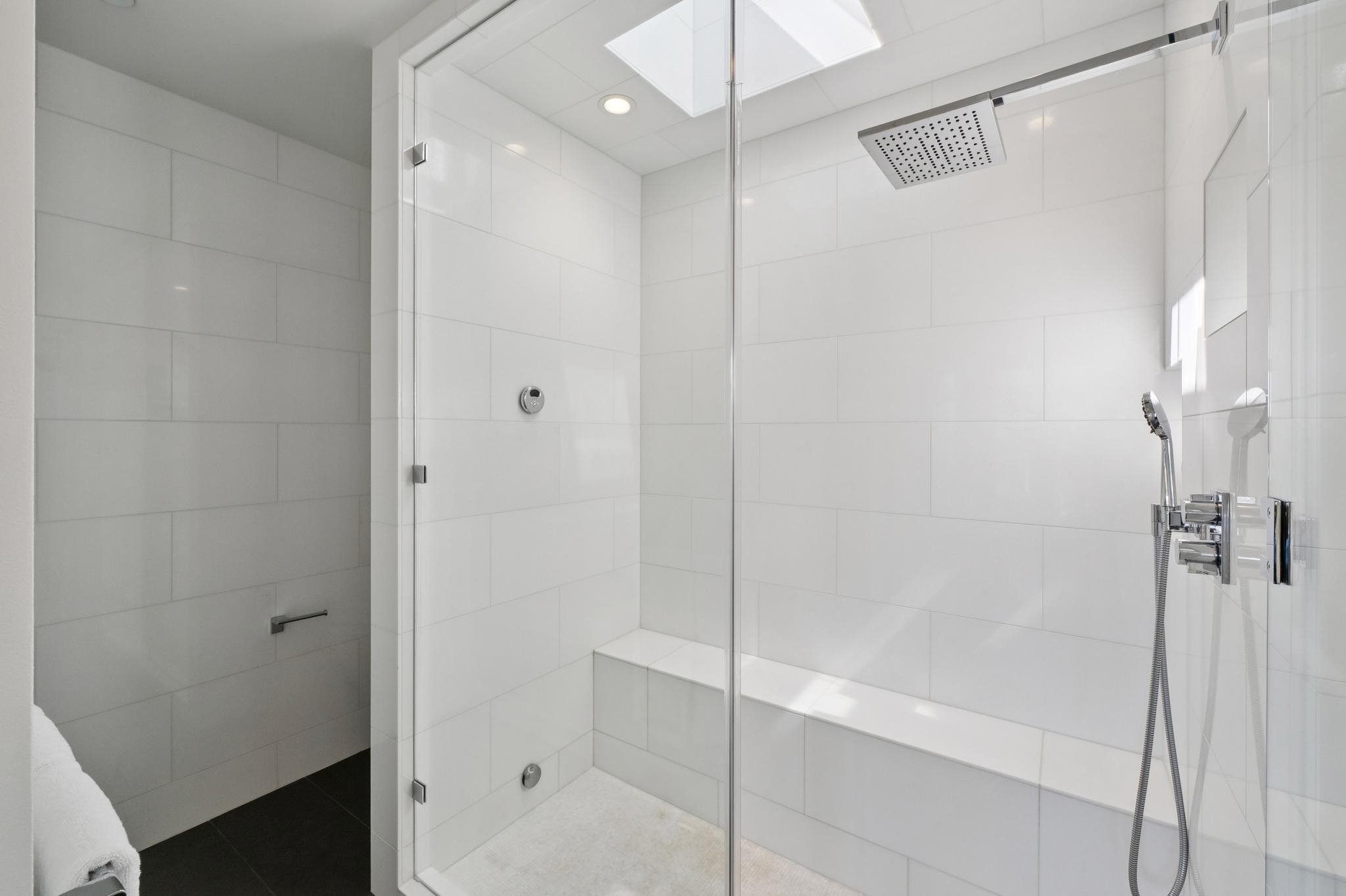 Modern white tiled walk-in shower with a glass door, overhead shower head, handheld shower wand, and a built-in bench.