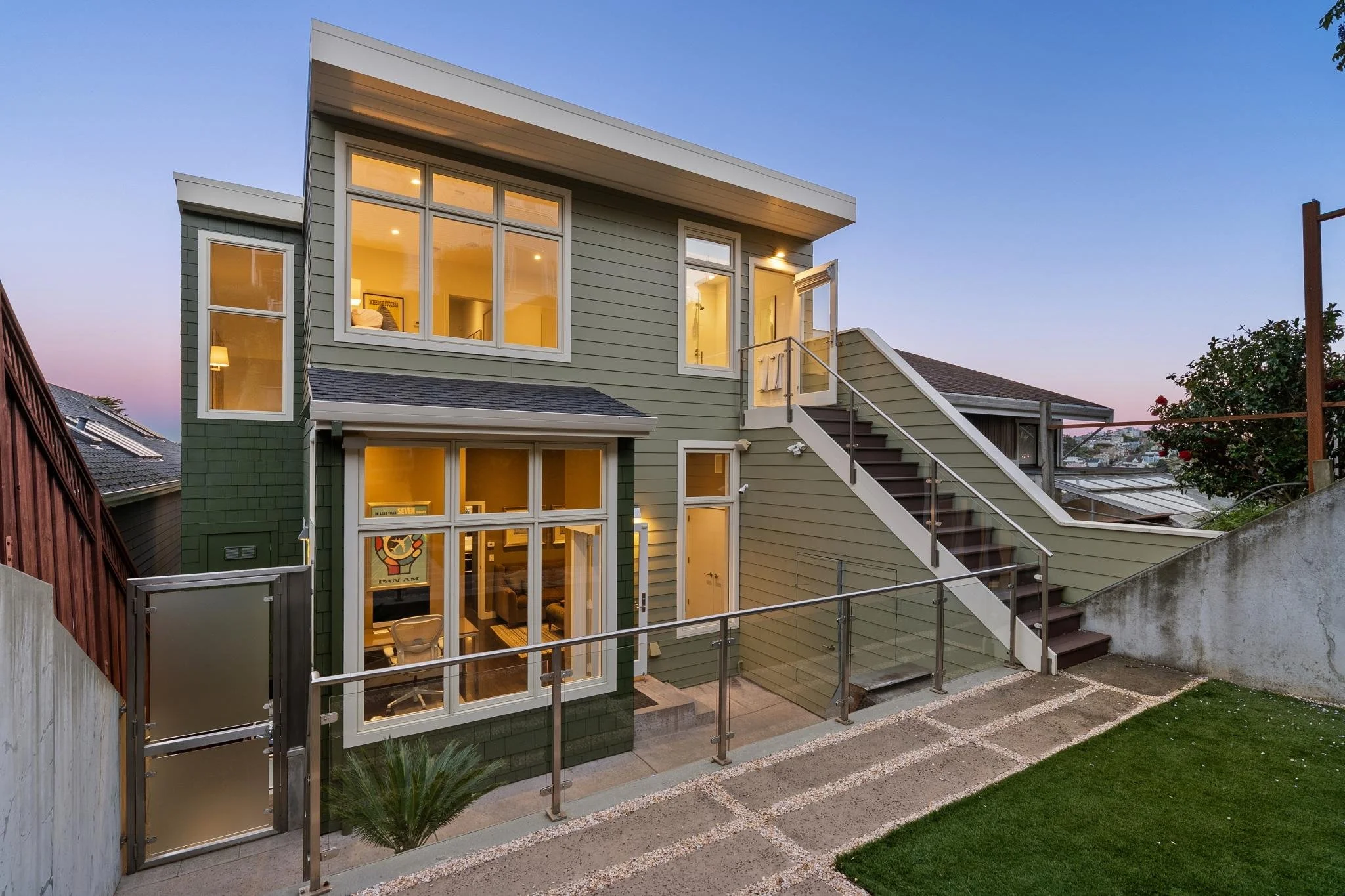 Modern three-story house during twilight with large windows, exterior stairs, a small gate, and a backyard with a grassy area and concrete walking path.
