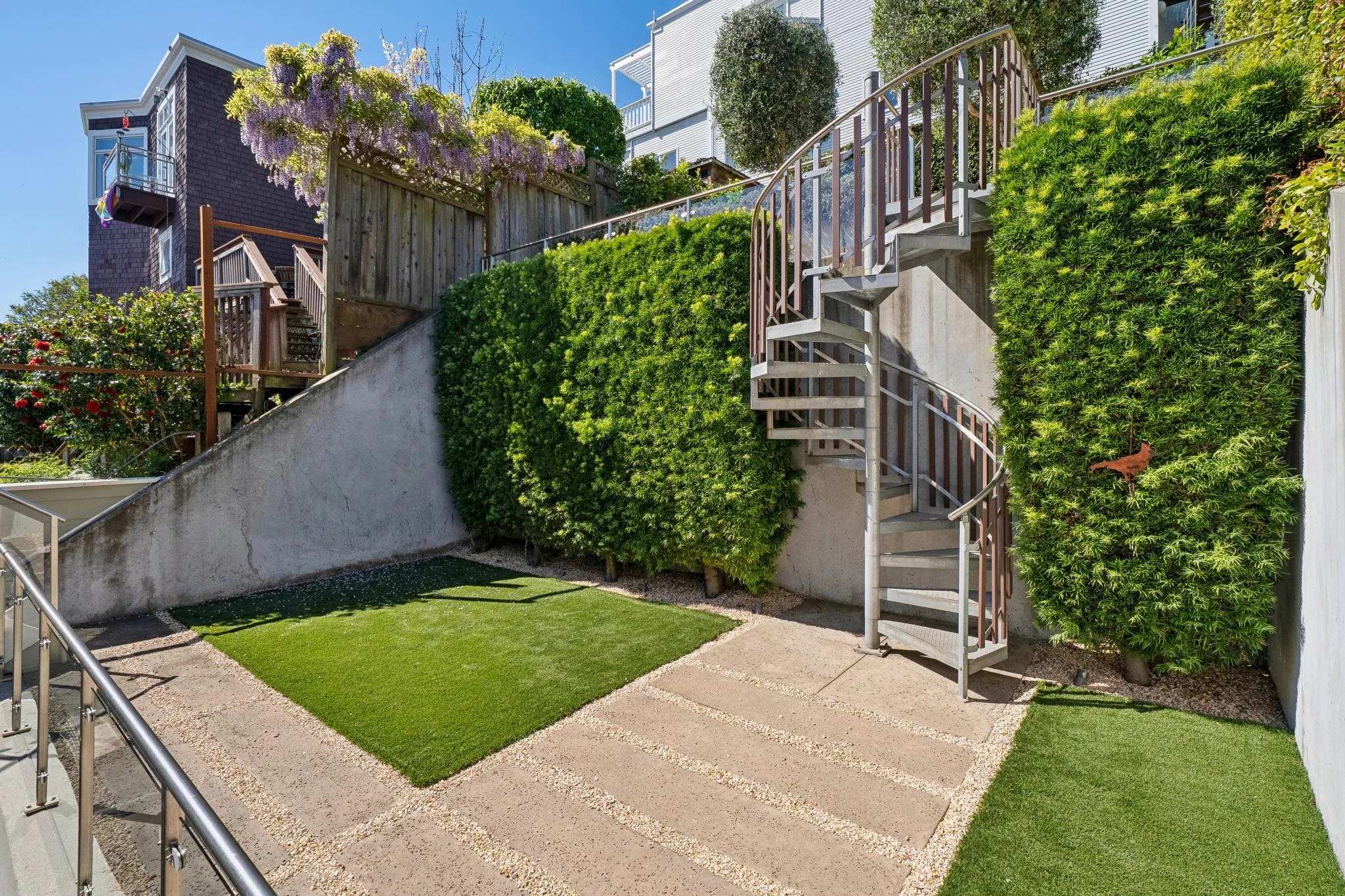 Small backyard with artificial grass, a concrete patio, and a metal spiral staircase leading to an upper level. The yard is bordered by green bushes and a concrete wall, with houses in the background.