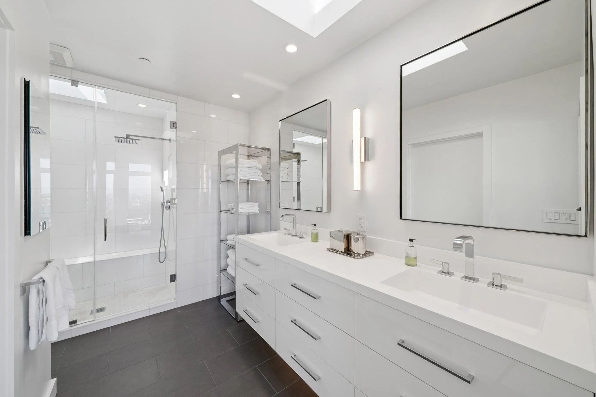 Modern white bathroom with double sink vanity, large mirrors, and a glass-enclosed shower with a rainfall showerhead. Towel rack and shelves with neatly folded towels.