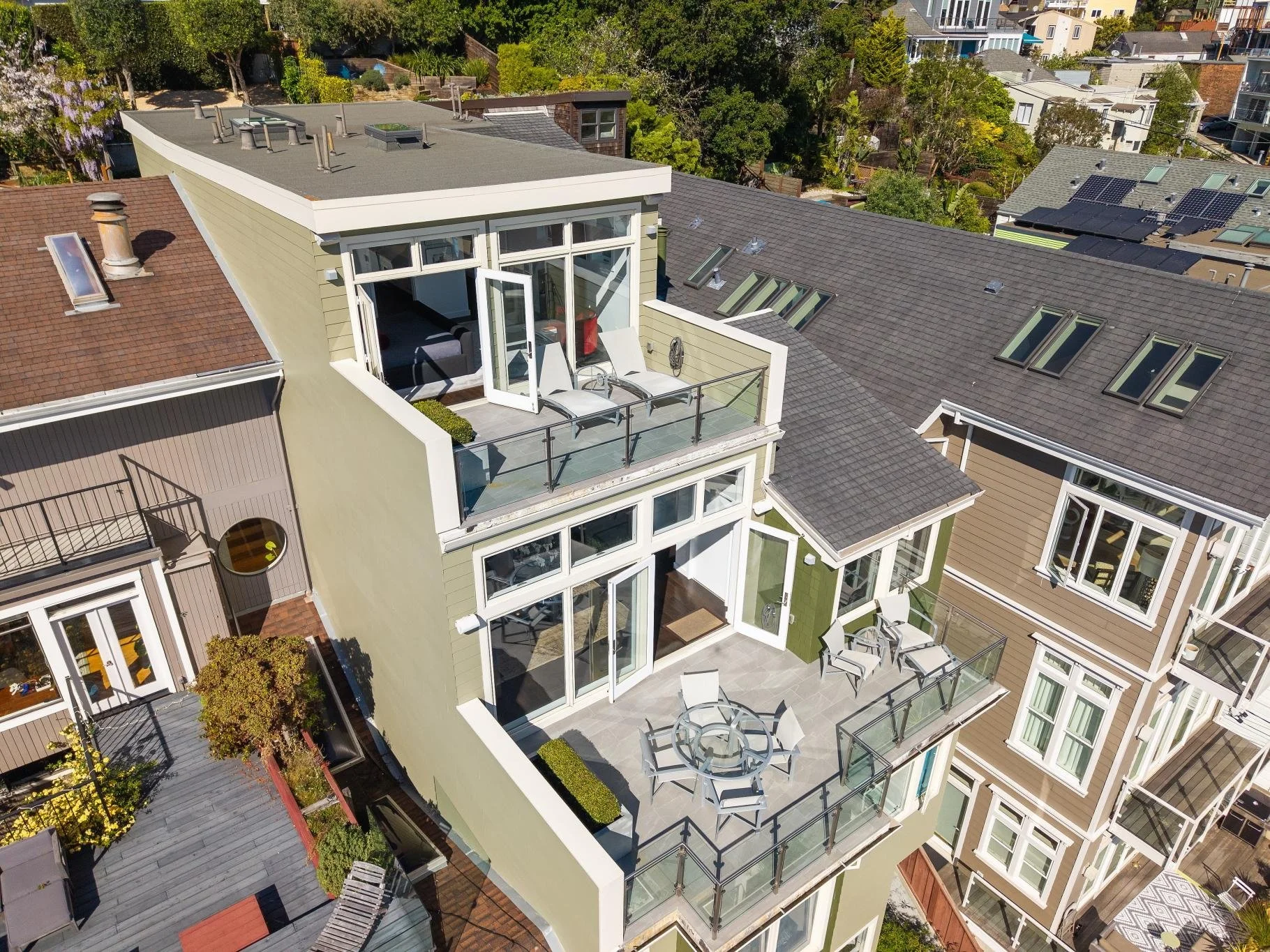 An aerial view of a modern apartment building with balconies, outdoor furniture, and patio areas, with neighboring rooftops and greenery in the background.