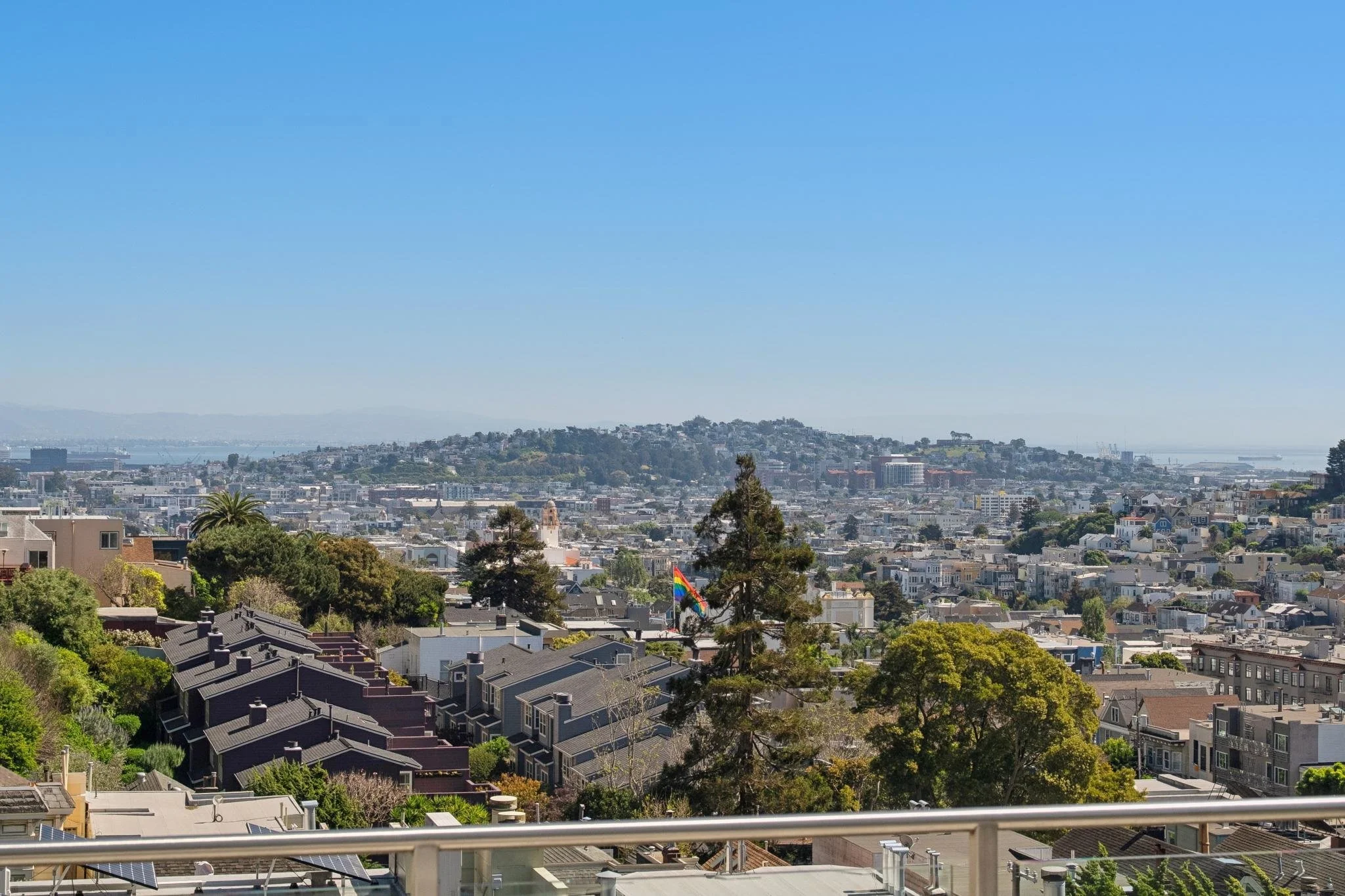 A cityscape with residential buildings, trees, and hills in the background, under a clear blue sky. A rainbow pride flag is visible among the trees.