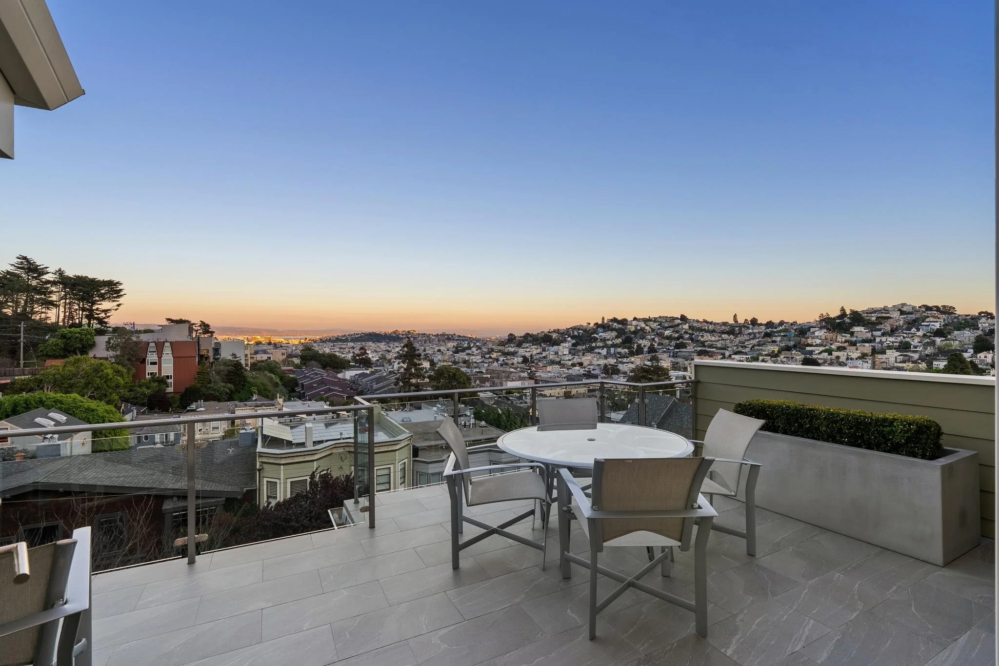 Balcony with a round table and four chairs overlooking a hilly neighborhood at sunset.