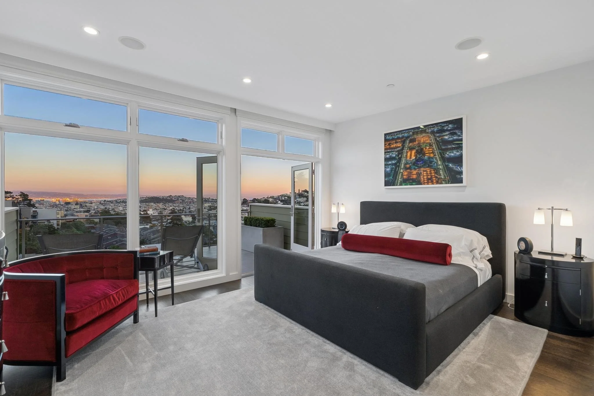 Modern bedroom with large windows showing city skyline at sunset, queen bed with dark gray headboard, red bolster pillow, nightstands with lamps, red velvet armchair with black frame, balcony with seating, contemporary art on white wall, soft gray carpet.