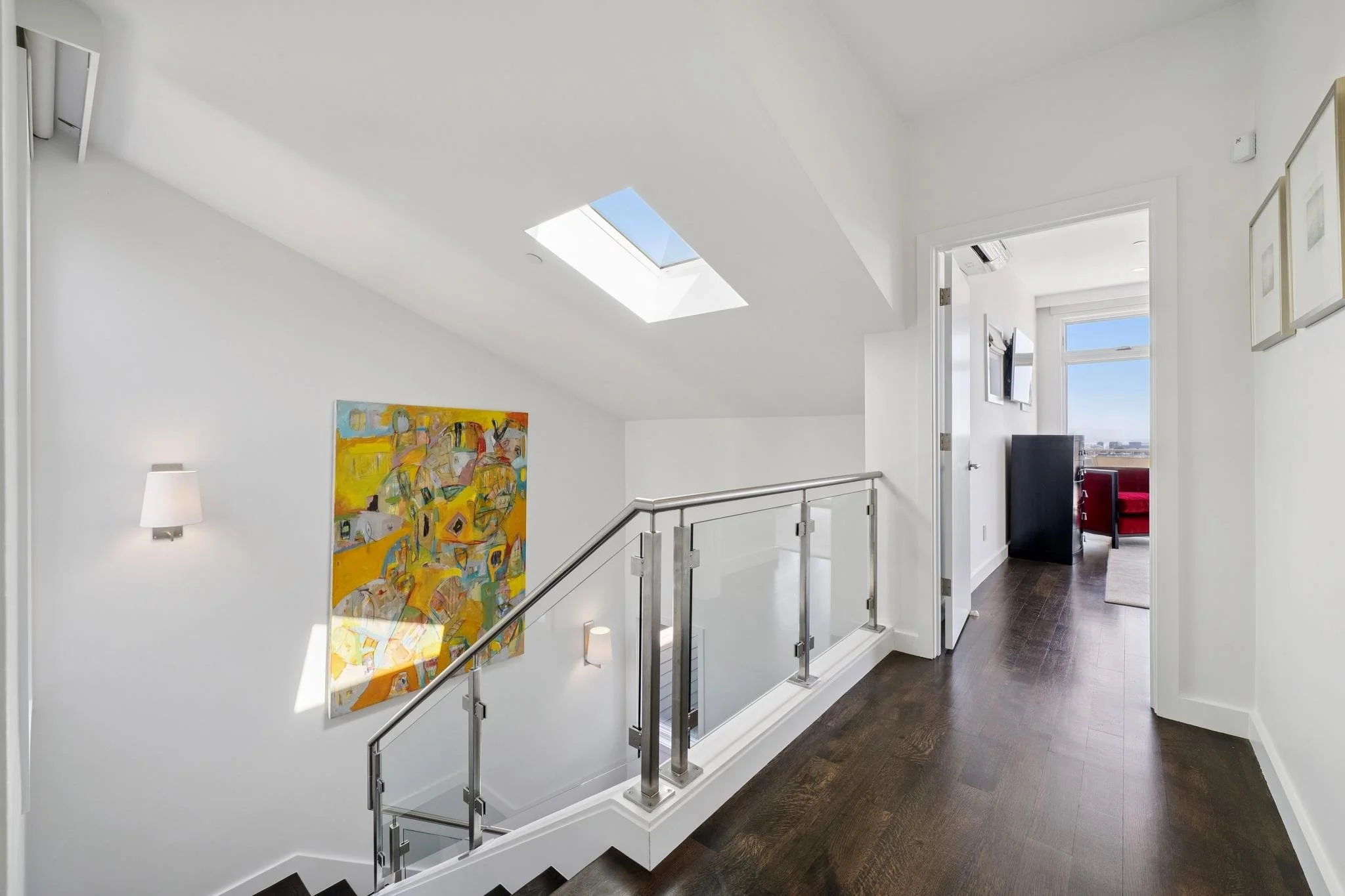 Bright hallway with white walls and dark hardwood floors, featuring a skylight and colorful abstract artwork, leading to a room with a window and red furniture.