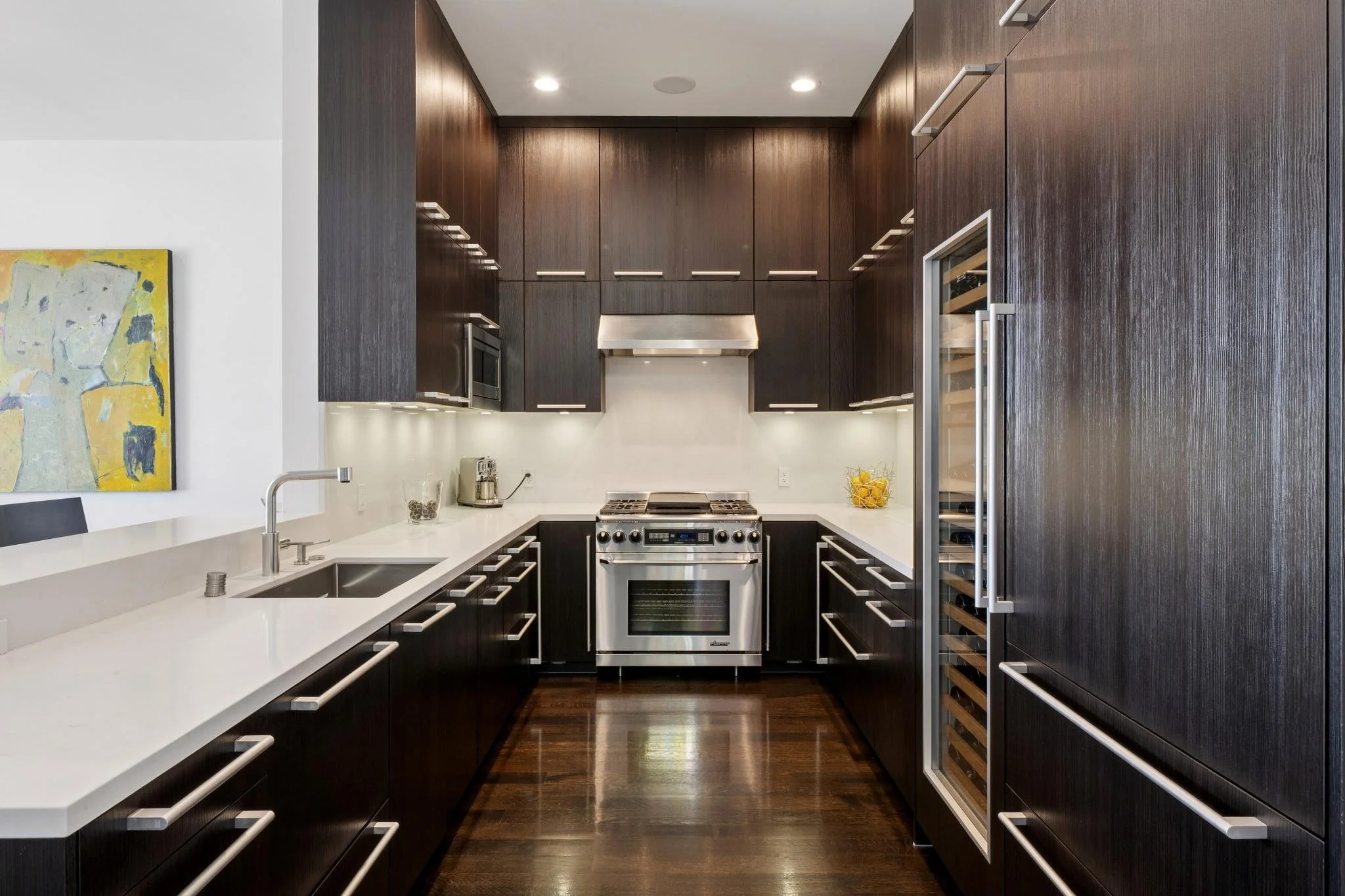 Modern kitchen with dark wood cabinets, white countertops, stainless steel appliances including a stove, microwave, and wine fridge, and hardwood flooring.