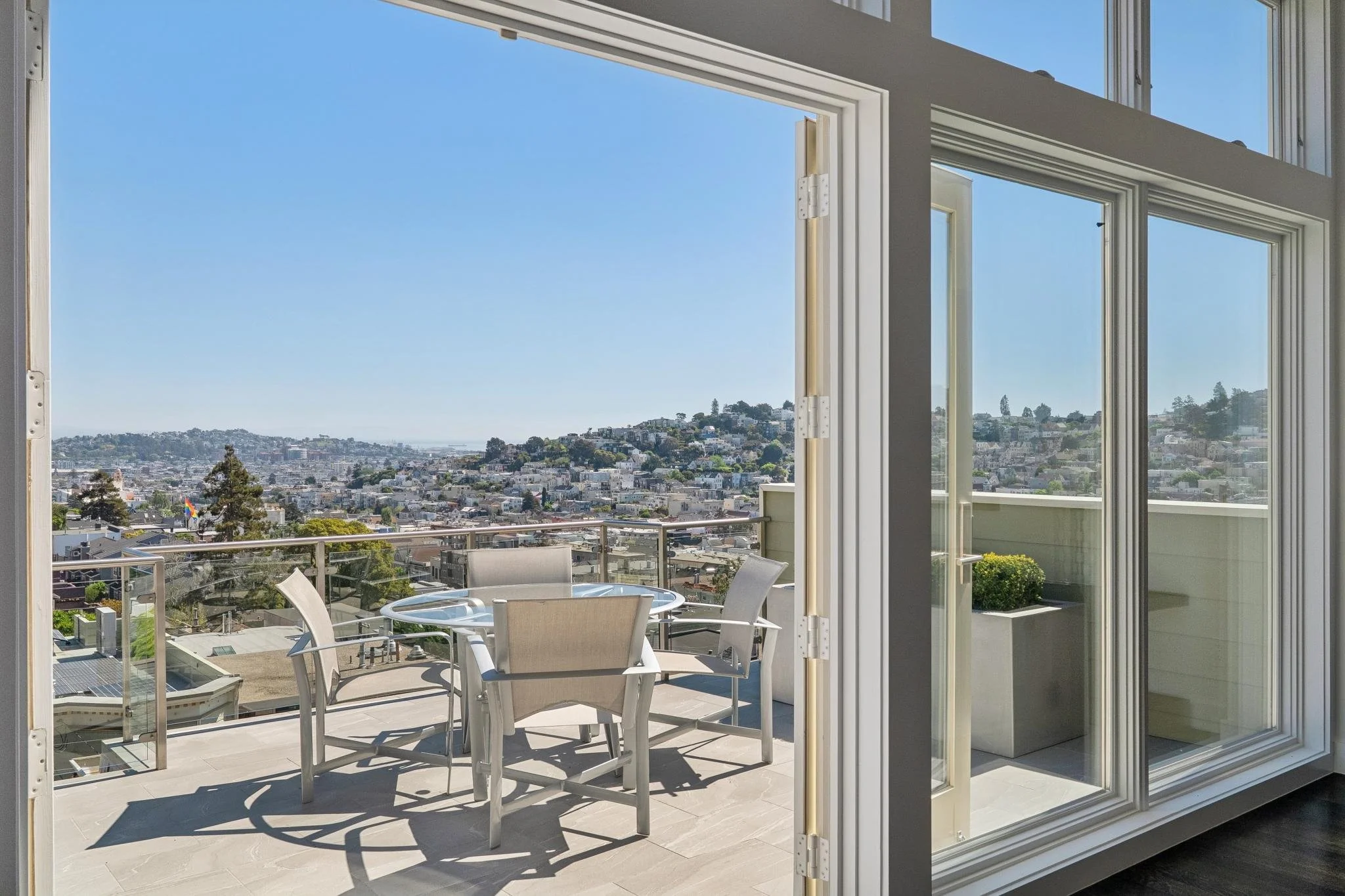 Balcony with a round glass table and four beige chairs, overlooking a cityscape on a clear day.