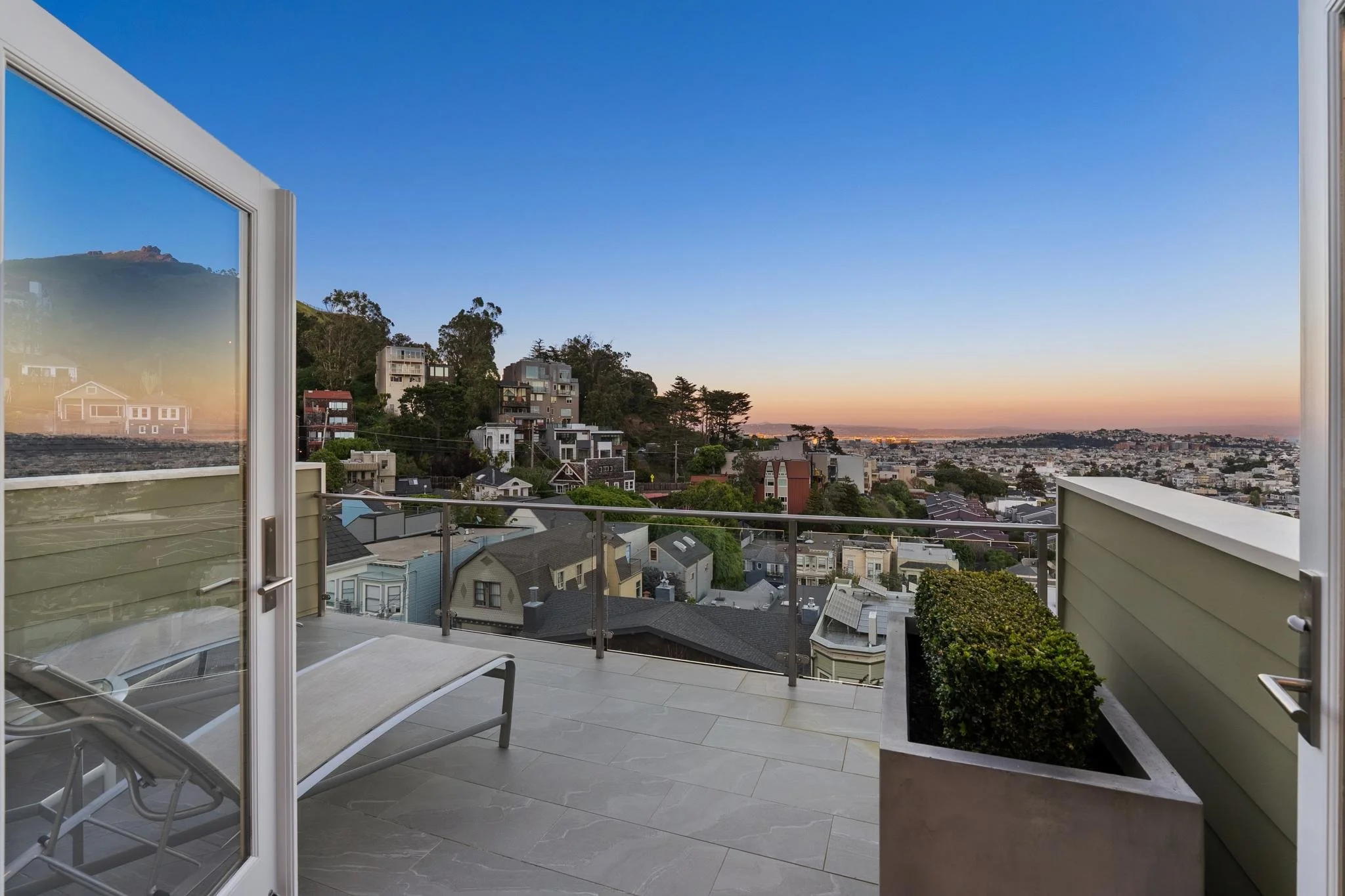 Balcony with a lounge chair, glass railing, planter with greenery, overlooking a cityscape with houses and trees at sunset.