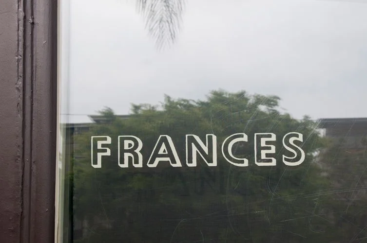 A glass window with the name 'FRANCES' written on it in white letters. Trees are reflected in the glass.