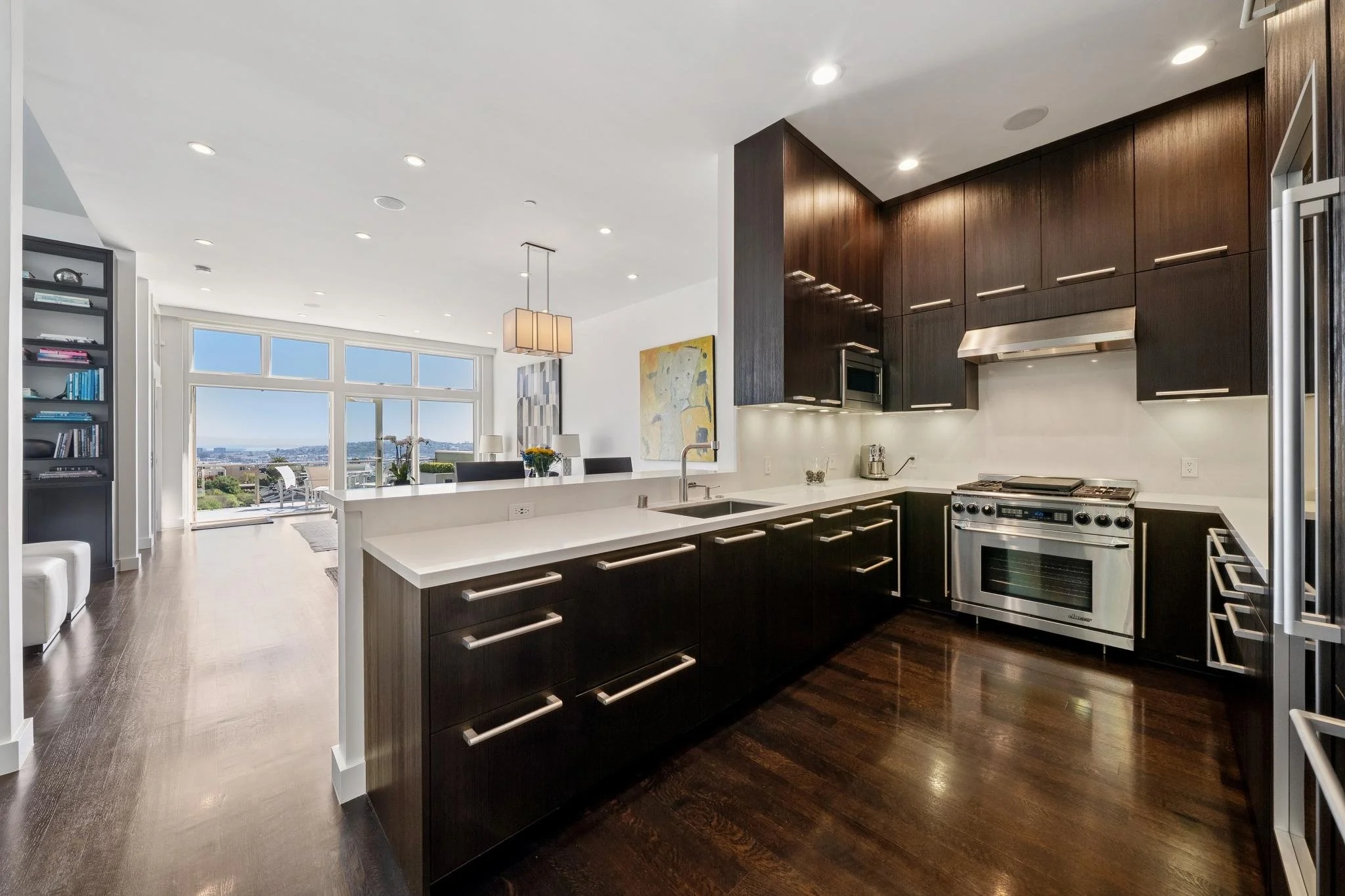 Modern kitchen with dark wood cabinets, white countertops, stainless steel oven and range, overlooking a bright living area with large windows and a city view.
