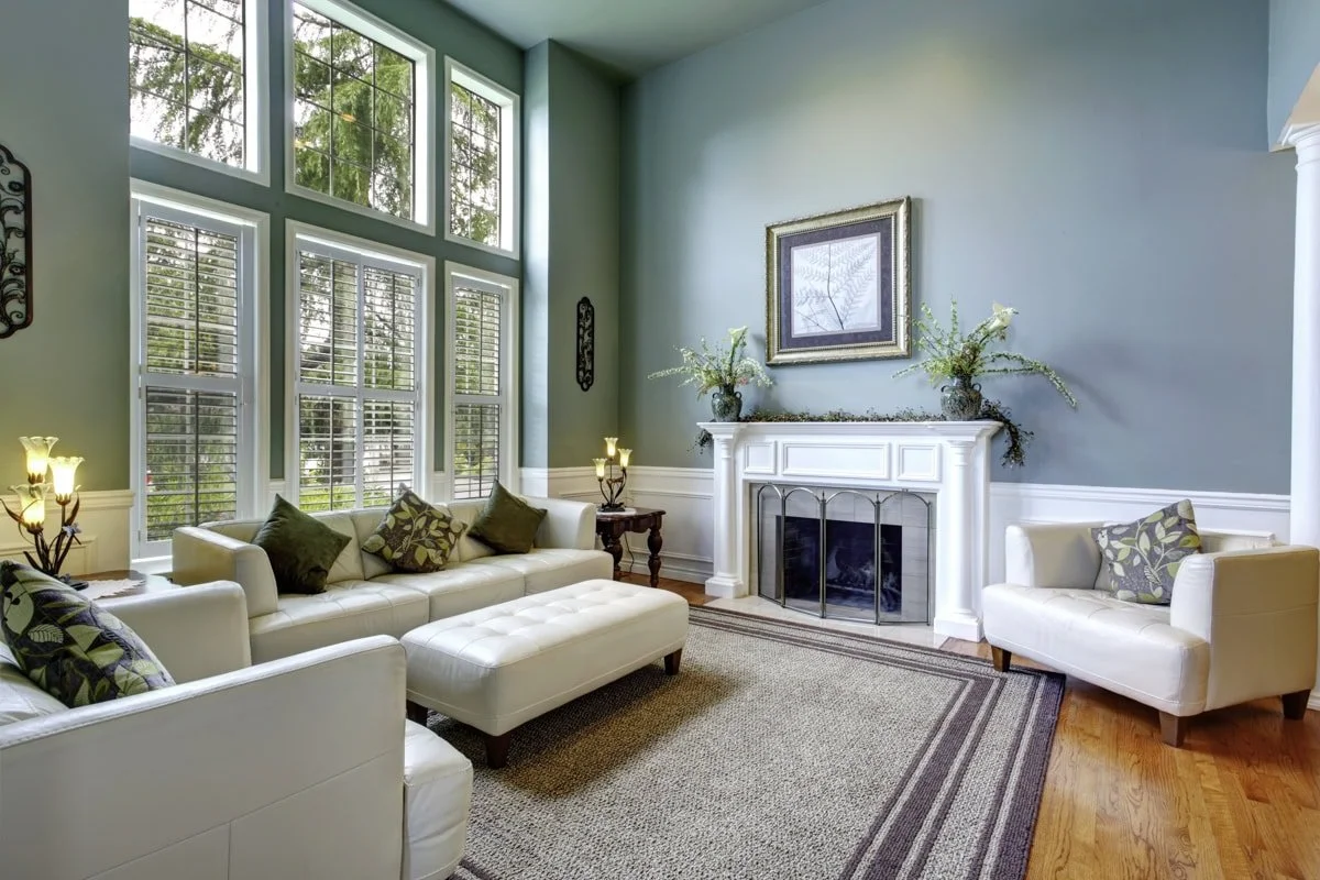 Living room with large windows, white leather sofa, armchair, and ottoman, fireplace with decorative vases and framed art above, hardwood floors, and area rug.