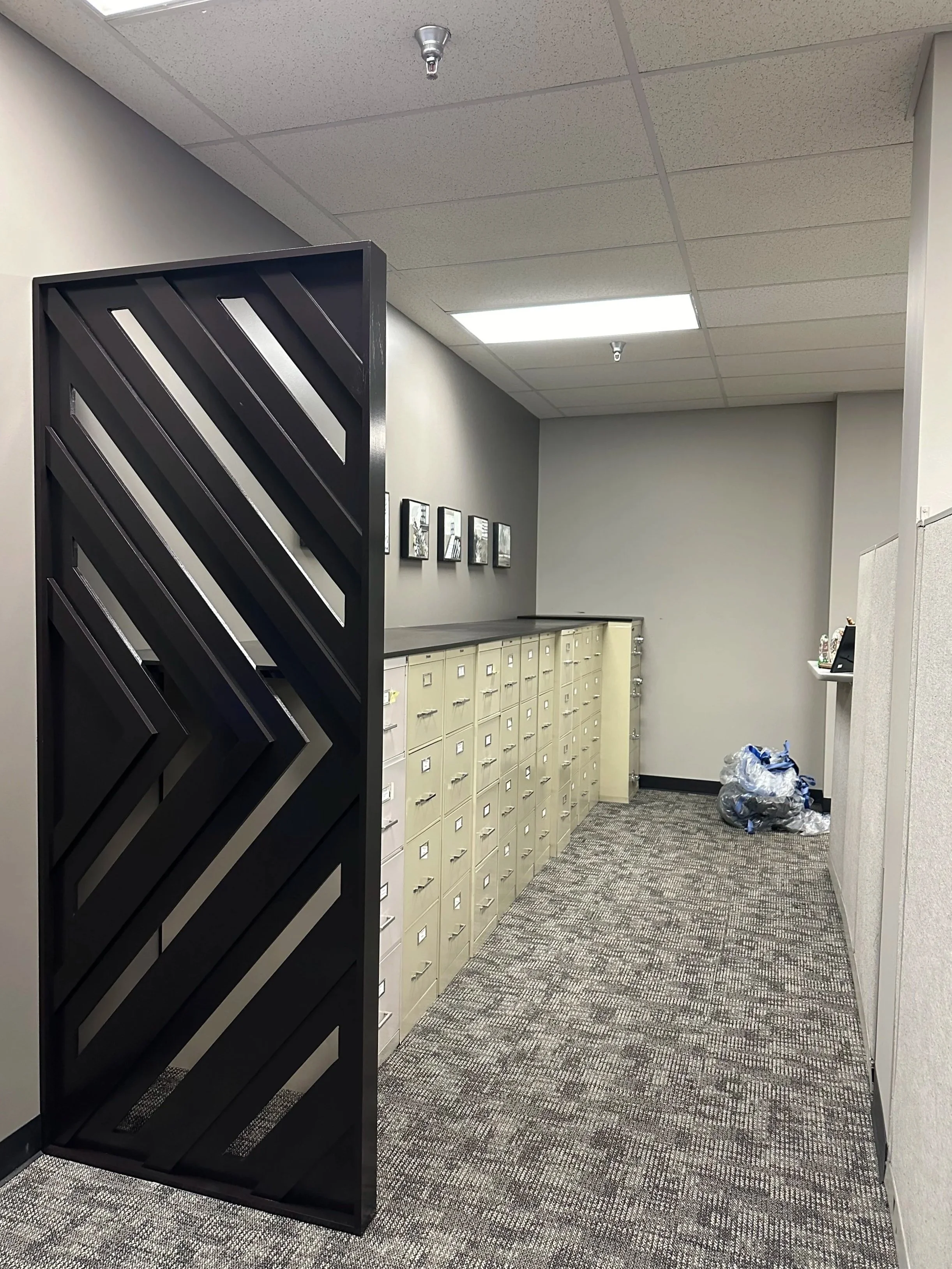 An interior office corridor with gray walls, beige lockers, and a black slatted room divider. Trash bags are on the floor, and there are framed pictures on the wall.