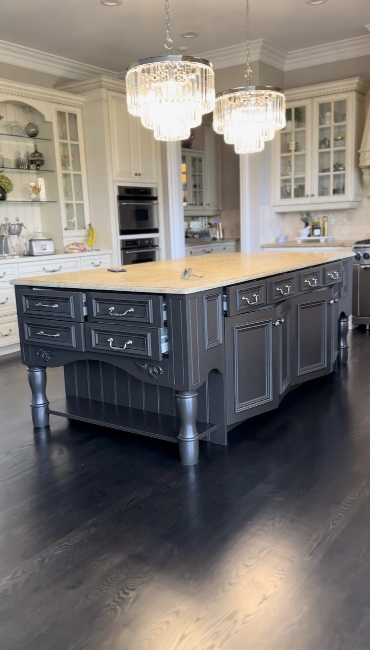 A kitchen with two elegant crystal chandeliers hanging above a large, dark gray kitchen island with a light granite countertop. The background features white cabinets with glass doors, built-in ovens, and various kitchen items.