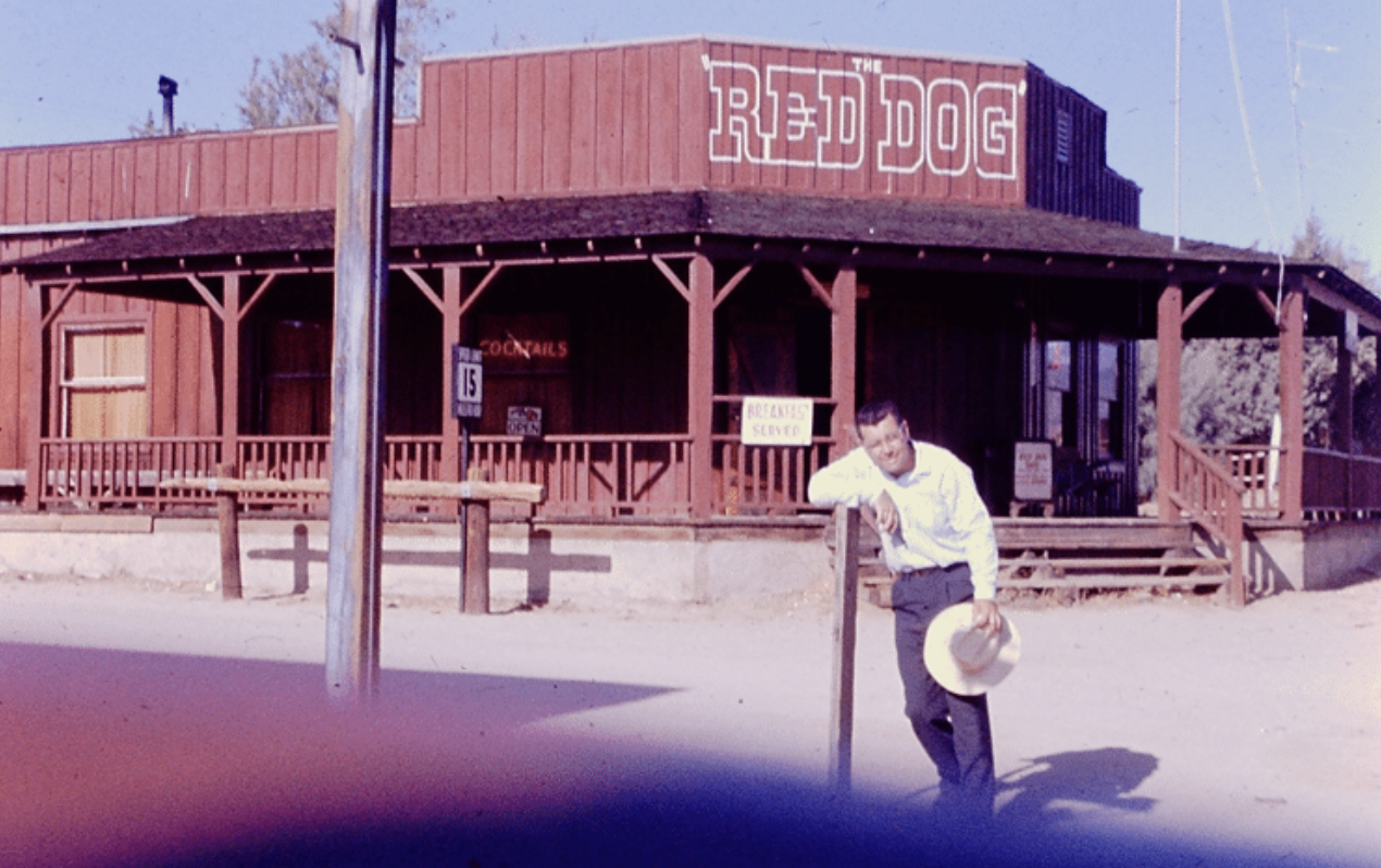 A man in a white shirt holding a hat leaning against a wooden post in front of a rustic wooden building named 'The Red Dog' with a porch, stairs, and signs advertising breakfast and drinks.