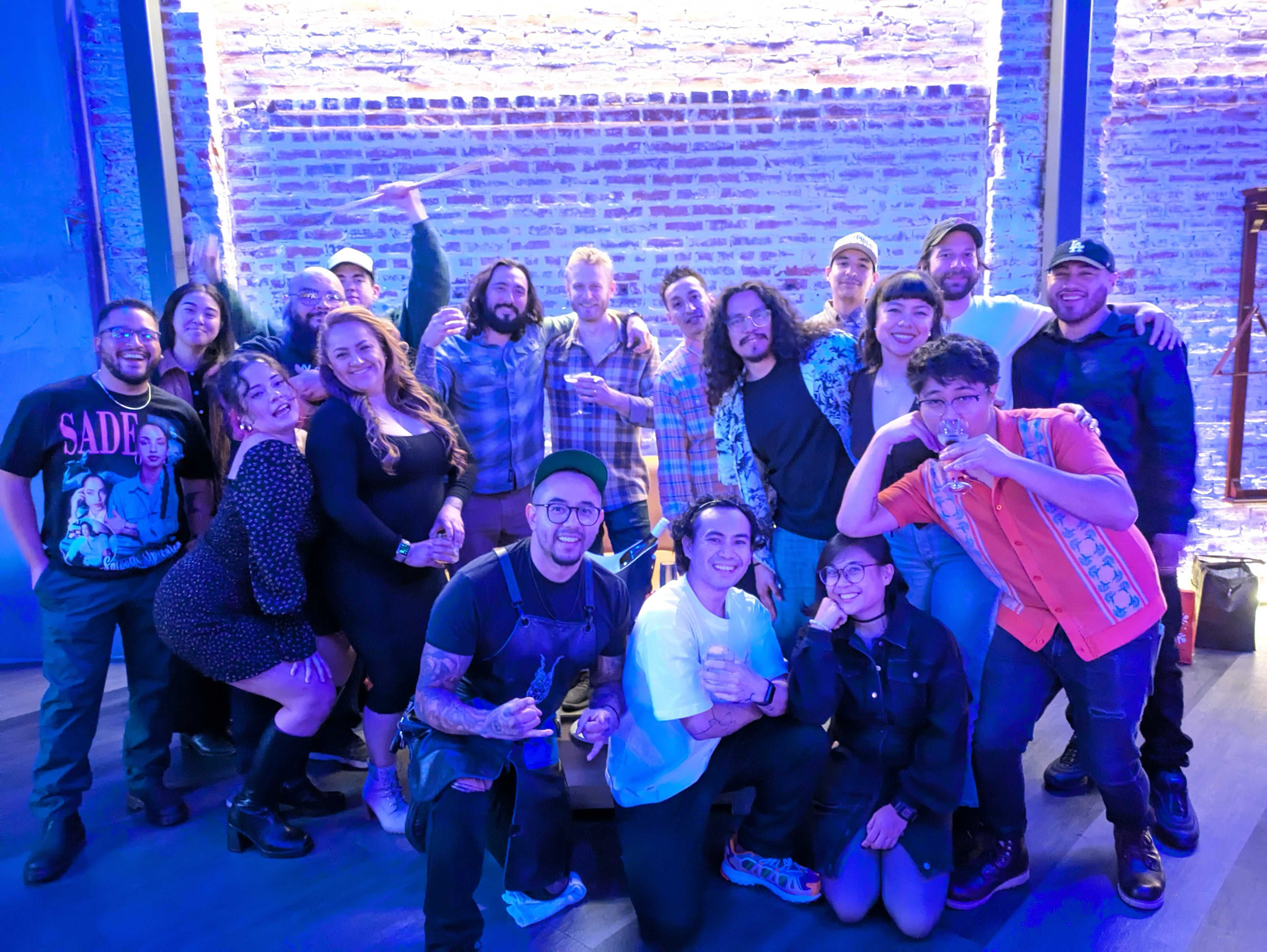 Group of people posing for a photo at a party or social gathering against a brick wall, some wearing casual clothes, holding drinks, smiling and making gestures.