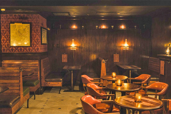 Cozy restaurant with wooden furniture, round tables, leather chairs, and warm wall lighting.