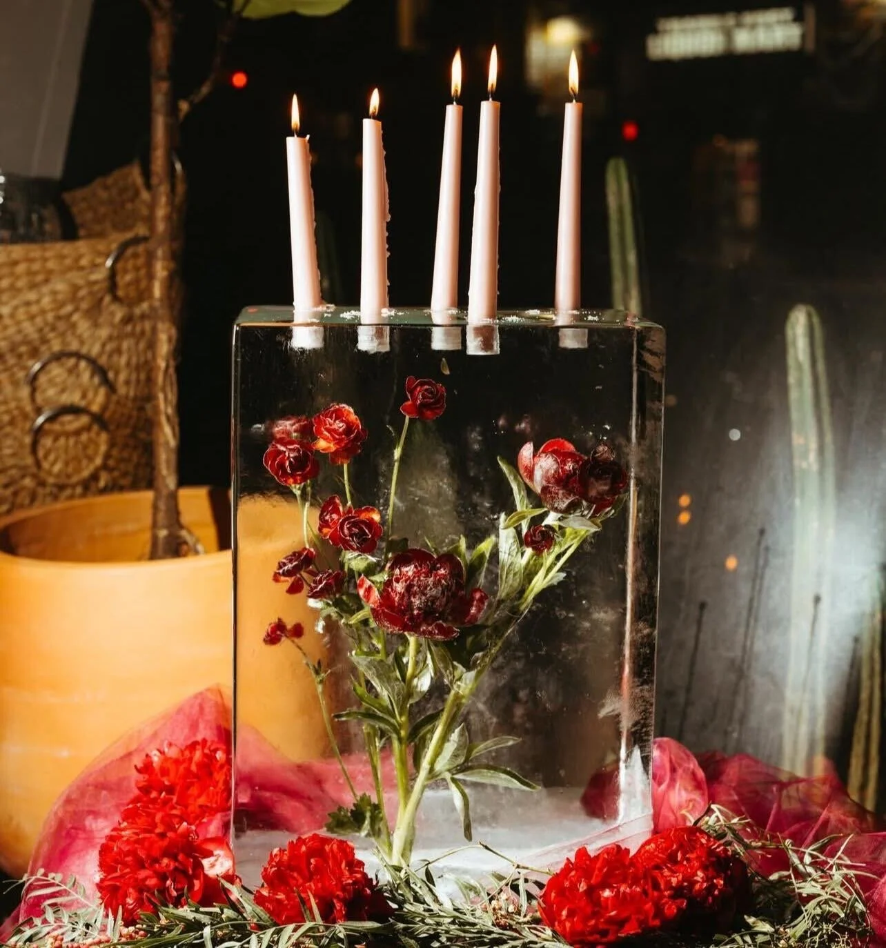 A glass vase with red flowers and five lit candles on top of the vase, surrounded by red flowers and greenery.