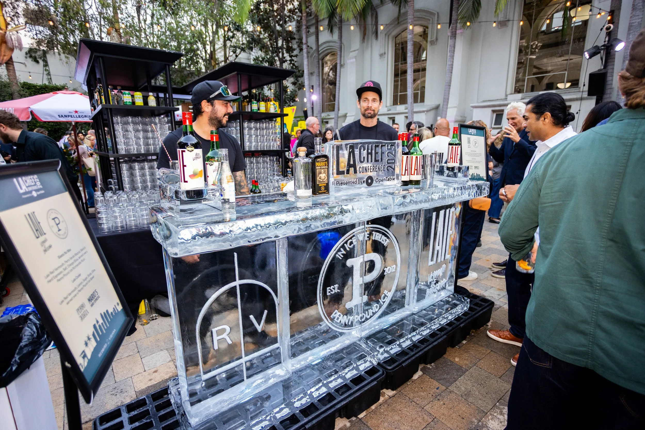 People gather around a bartender at a beverage stand with a clear ice bar at an outdoor event, with strings of lights hanging overhead and a historic building in the background.