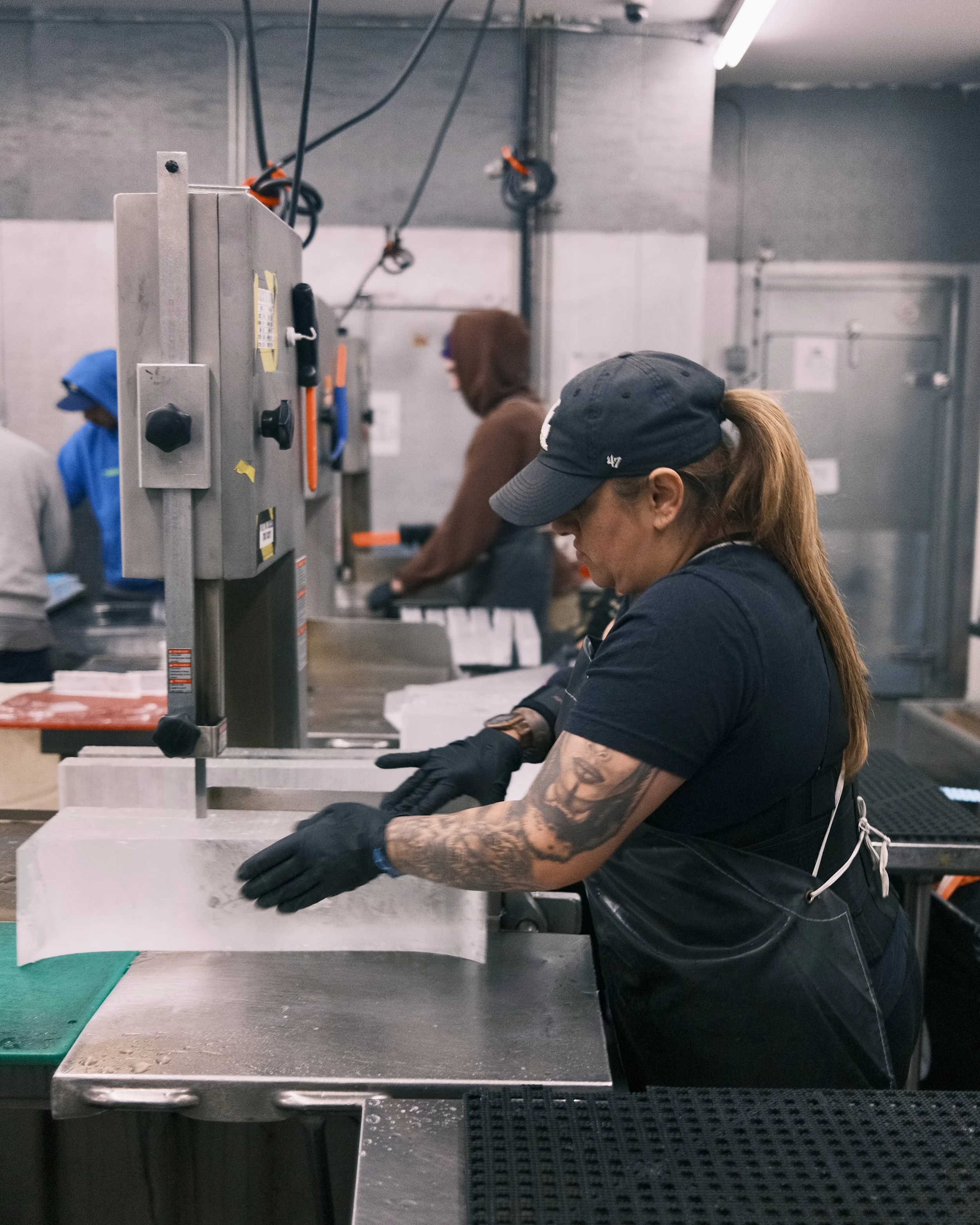 A woman with a ponytail and tattoos on her arm, wearing a cap and black gloves, working at a metal work station in an industrial setting. She is focused on a task with her hands near a white block, possibly applying a process or detail to it.