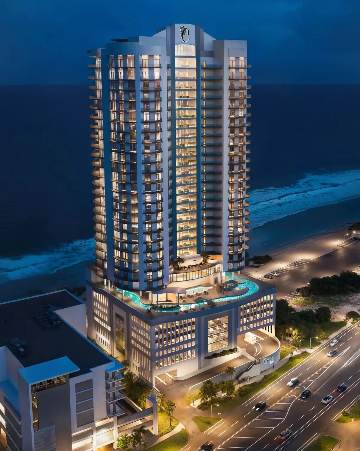 A tall modern hotel building at night with illuminated balconies and a rooftop pool, located near the beach with waves visible in the background.