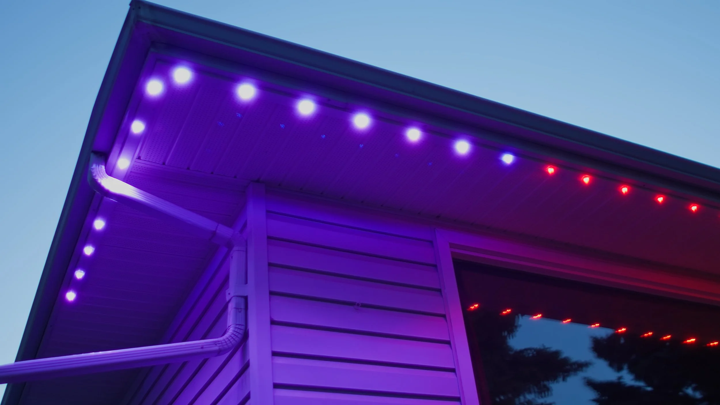 Close-up of a house with purple and red LED strip lights along the eaves at dusk, with trees visible in the background.