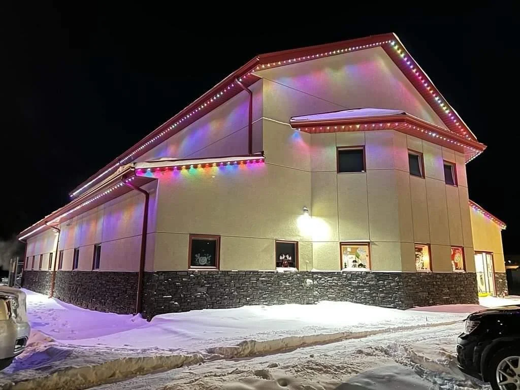 Building decorated with colorful Christmas lights at night, snow on the ground, parked cars in the foreground.