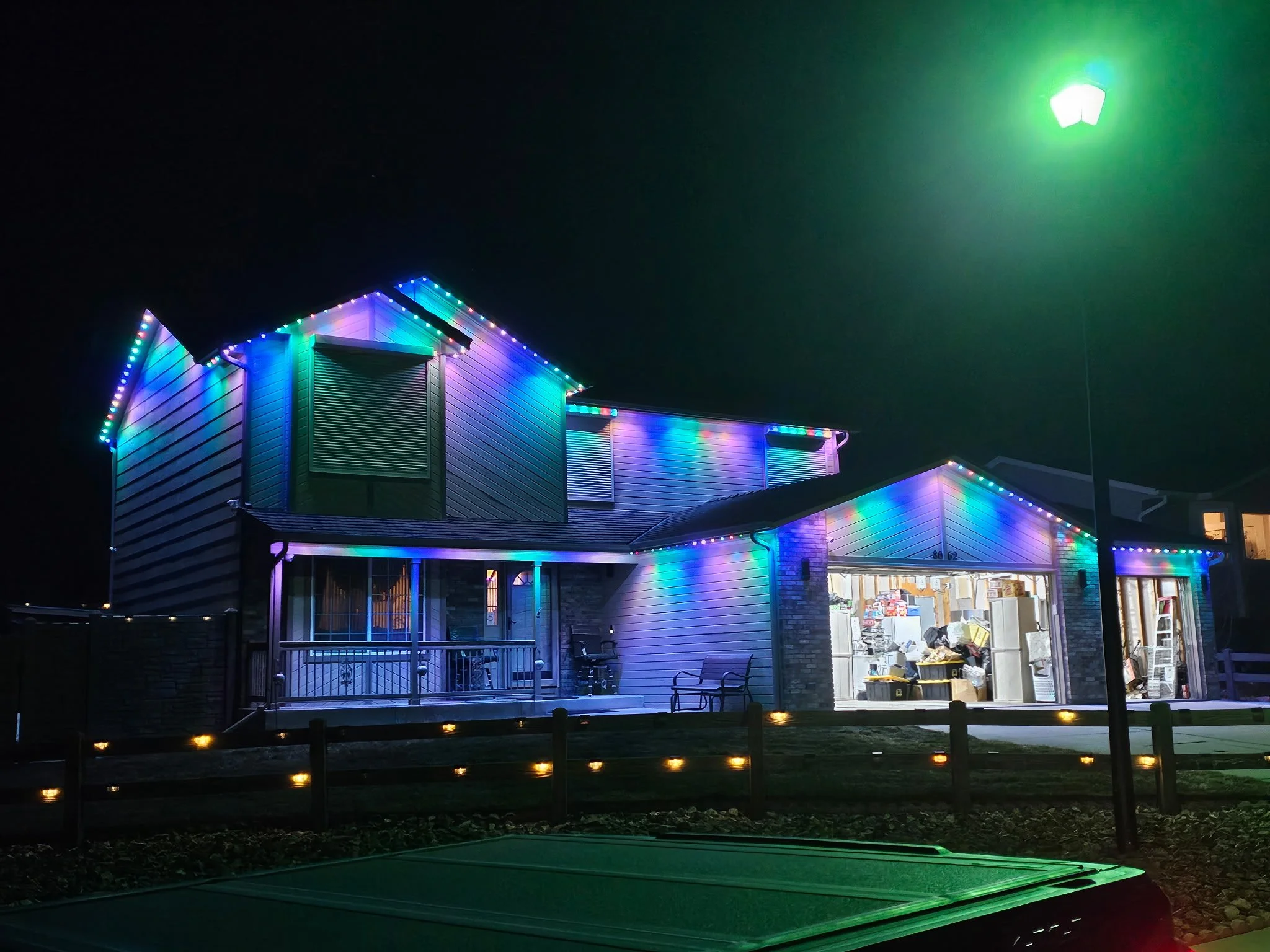 A house decorated with colorful outdoor Christmas lights at night, with a garage door open showing clutter inside, and a porch with outdoor furniture.