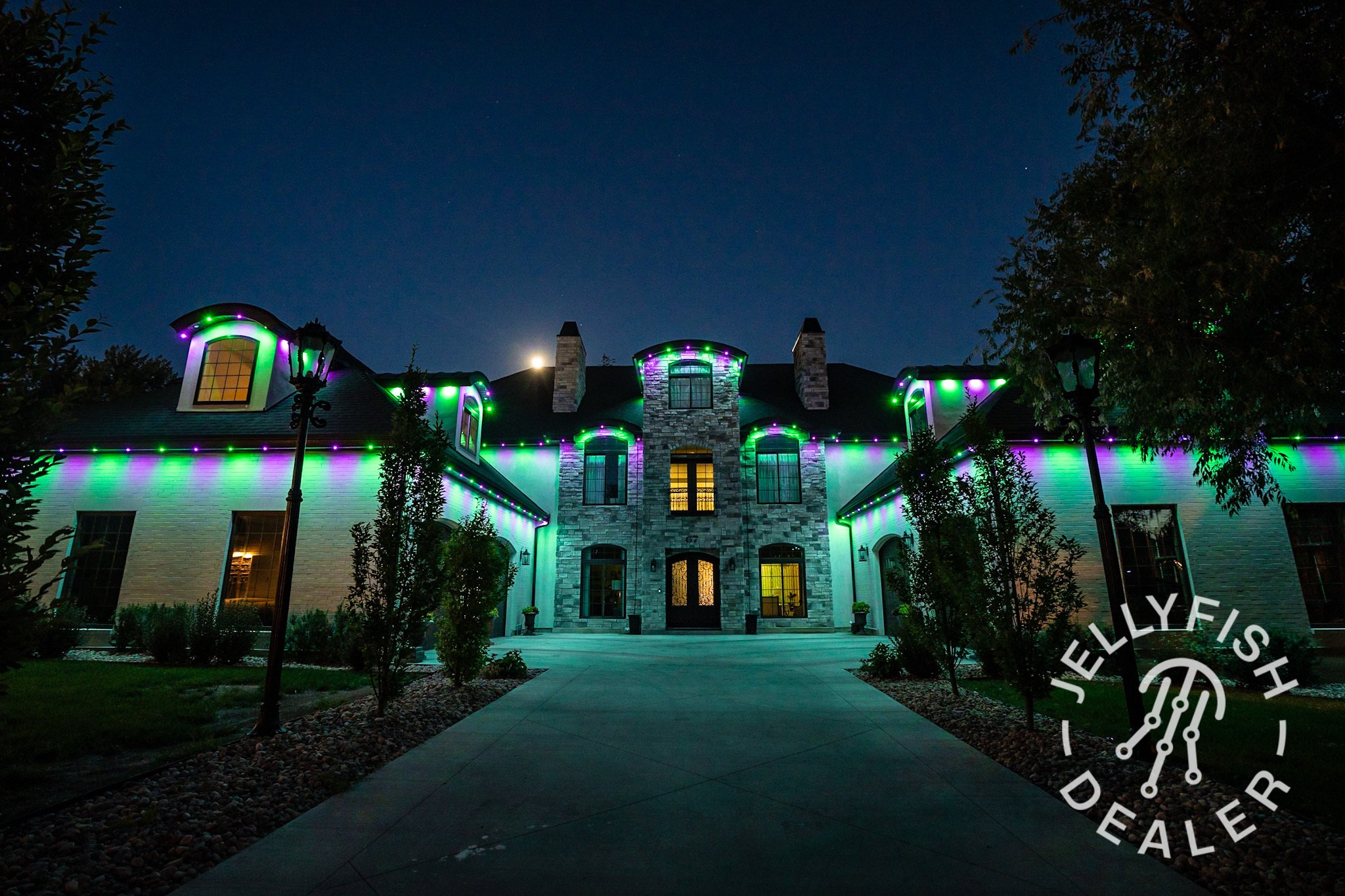 A large house illuminated with green and purple lights at night, with a paved walkway leading to the front door, surrounded by trees and street lamps.