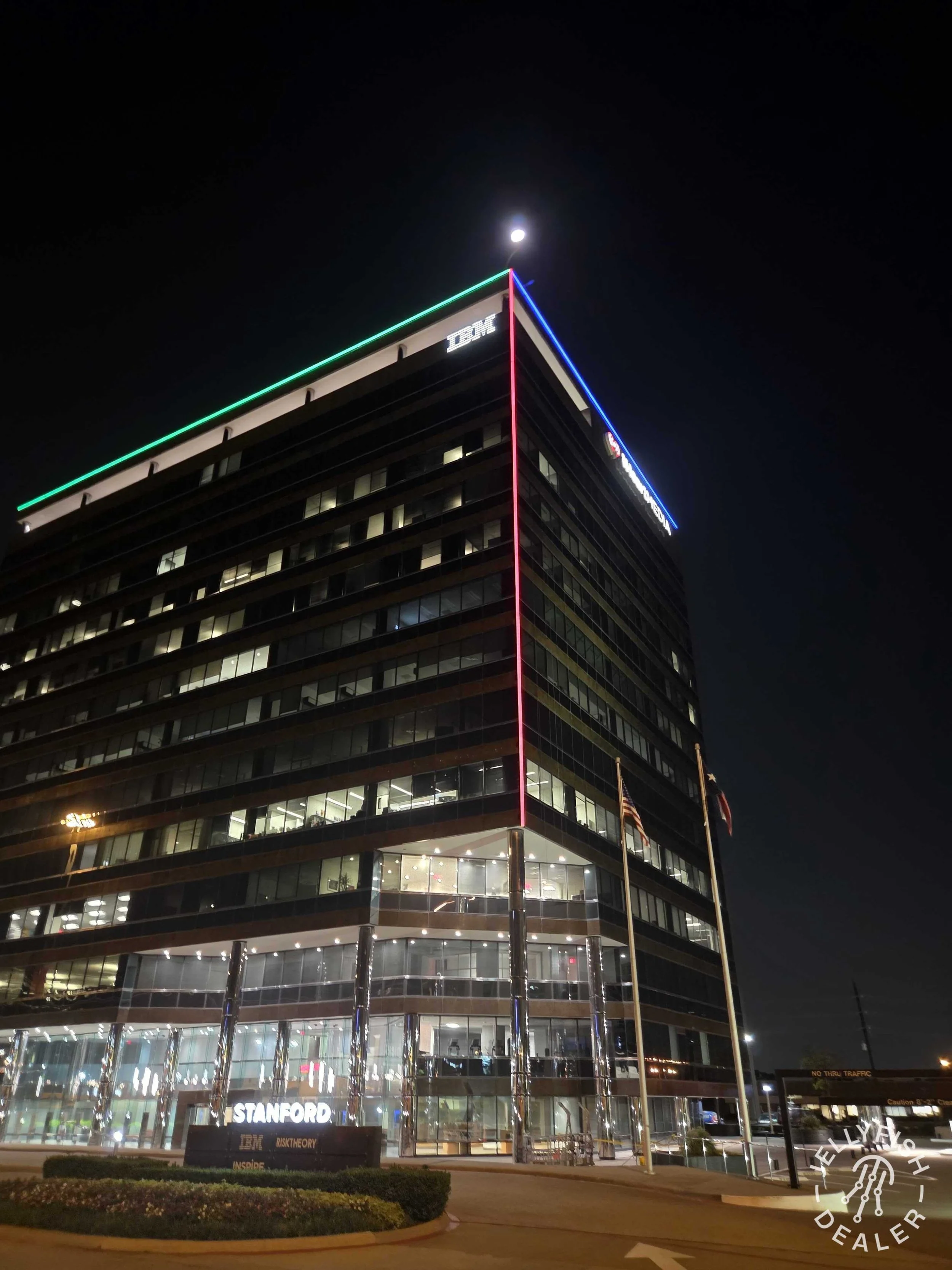 Night view of a multi-story office building with illuminated company logos for IBM and Stanford, neon lights along the rooflines, and a bright moon in the clear night sky.