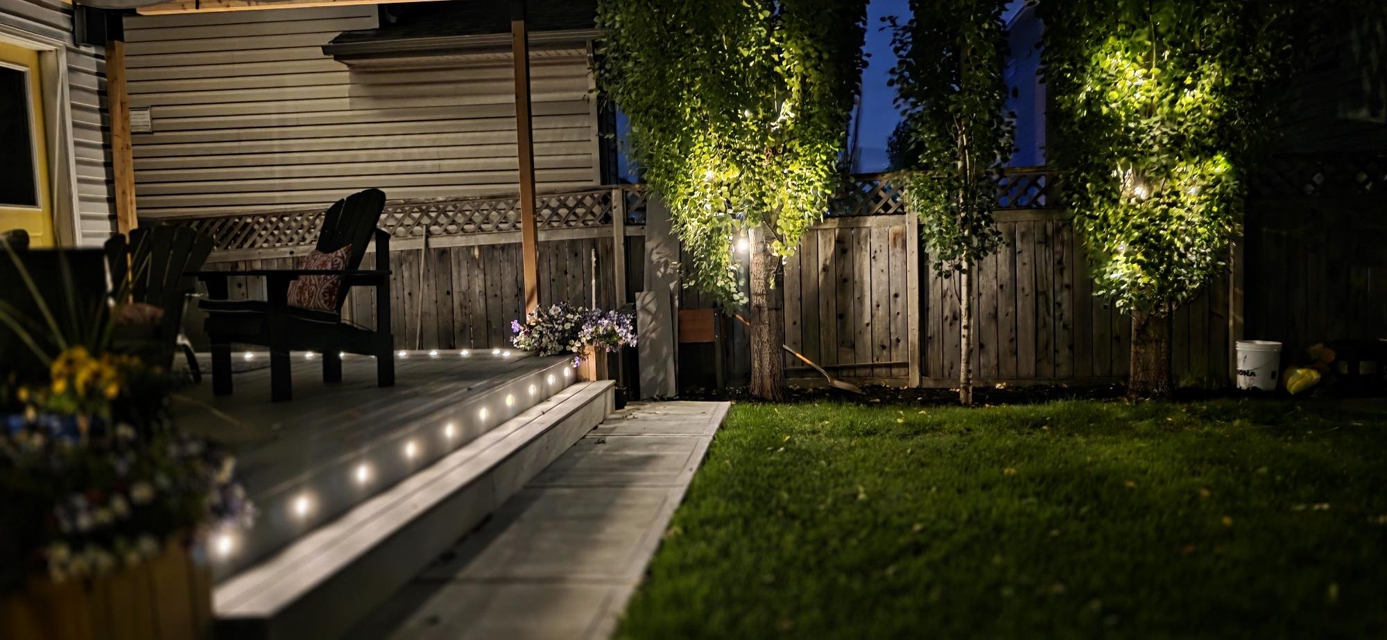 A backyard patio at night with string lights along the edge, a wooden deck with chairs, potted flowers, trees with lights, a grass lawn, and a wooden fence.