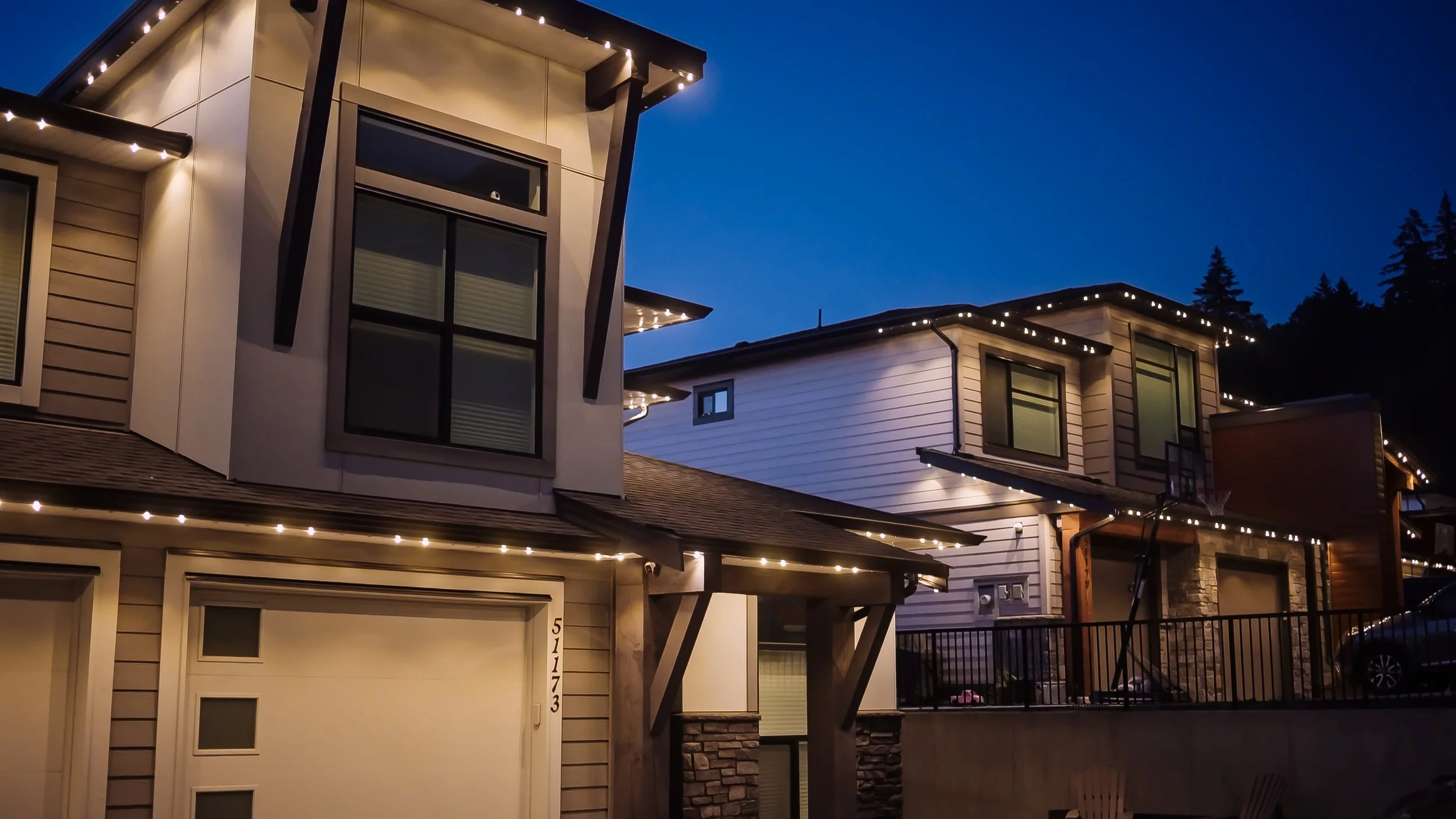 Modern multi-story houses decorated with string lights at dusk with a dark blue sky and trees in the background.