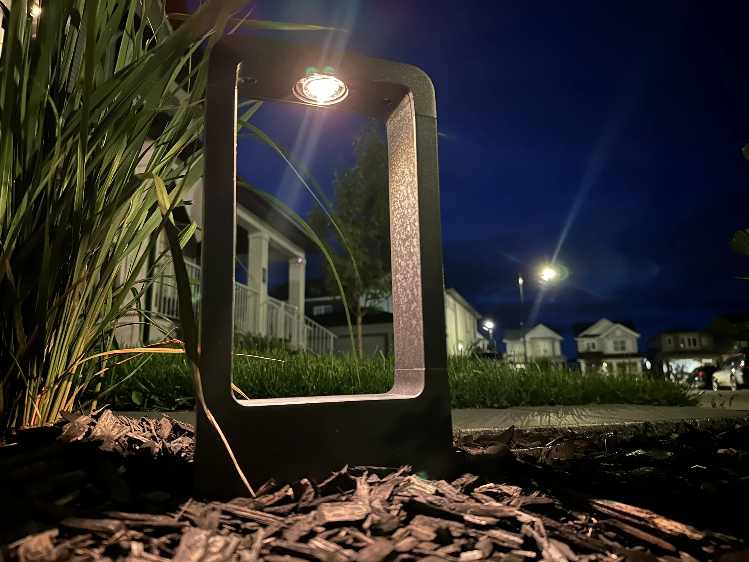 Close-up of a garden light fixture reflecting in a rectangular mirror on the ground at night, with surrounding grass, mulch, and in the background, houses and streetlights in a suburban neighborhood.