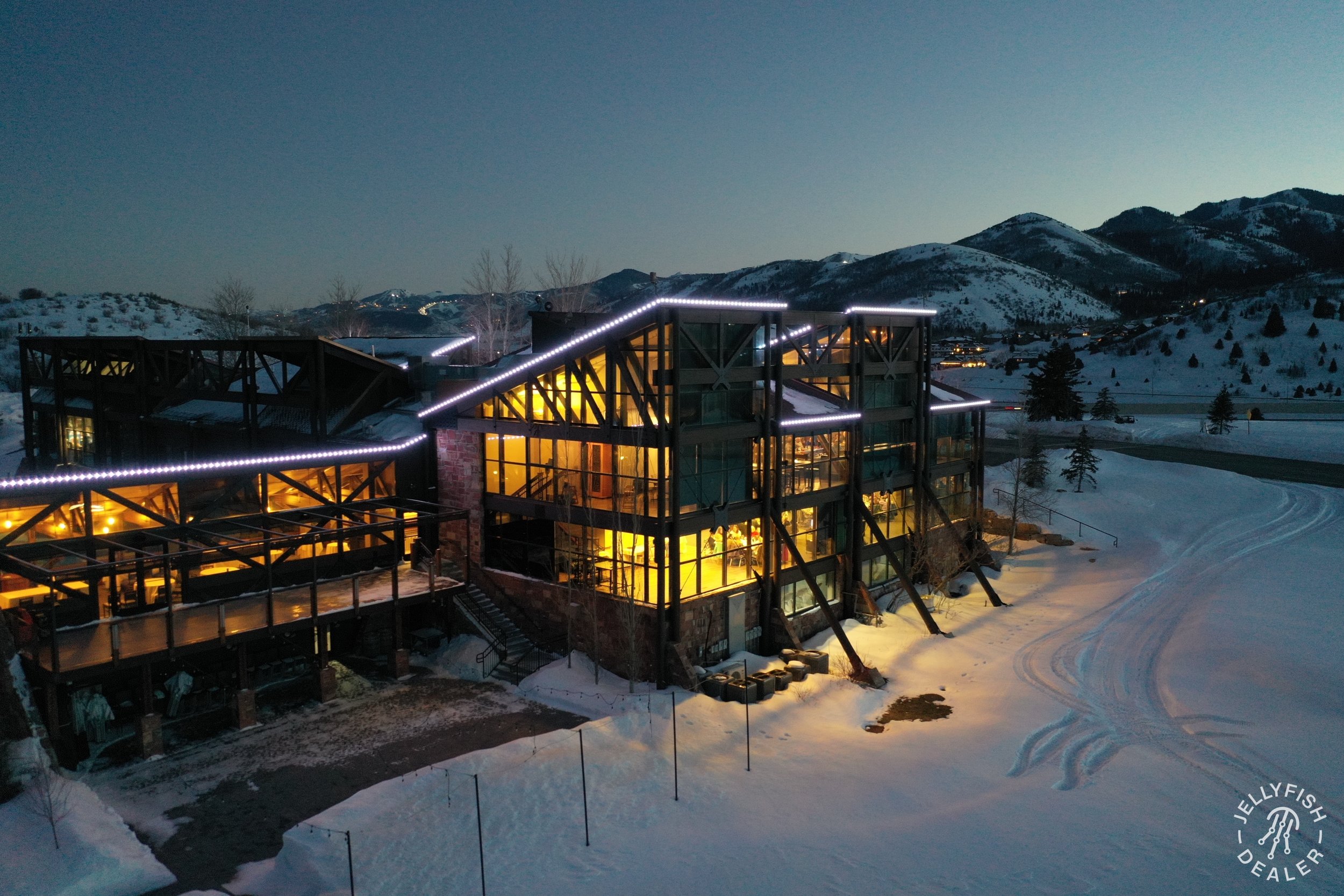 A modern glass and steel building with warm interior lighting, set against a snowy mountain landscape at dusk, with tracks in the snow nearby.