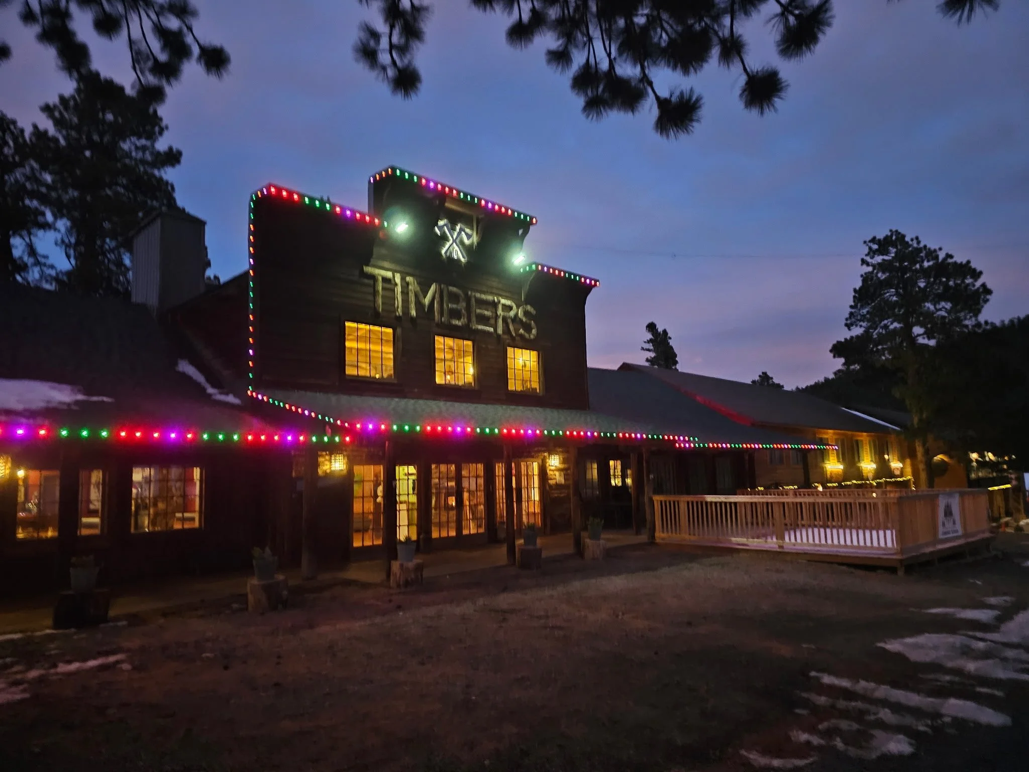 A rustic building with a sign that says 'TIMBERS' illuminated by colorful Christmas lights during dusk. The building has warm yellow lighting from the windows, a deck on the right side, and tall trees in the background.