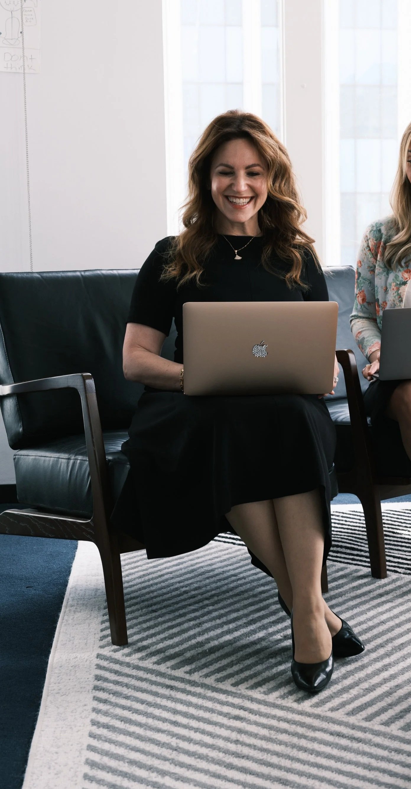 A woman with wavy brown hair, wearing a black dress and high heels, smiling while sitting on a black leather chair with a silver MacBook on her lap, in a well-lit room with large windows.