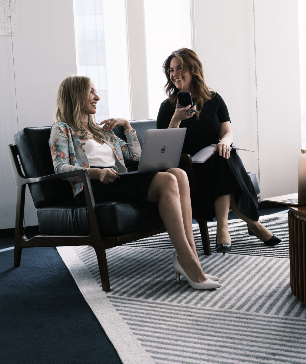Two women sitting on a black leather sofa, smiling and chatting in an office. One woman holds a laptop on her lap, the other holds a smartphone and a notebook. They are in a bright room with large windows.
